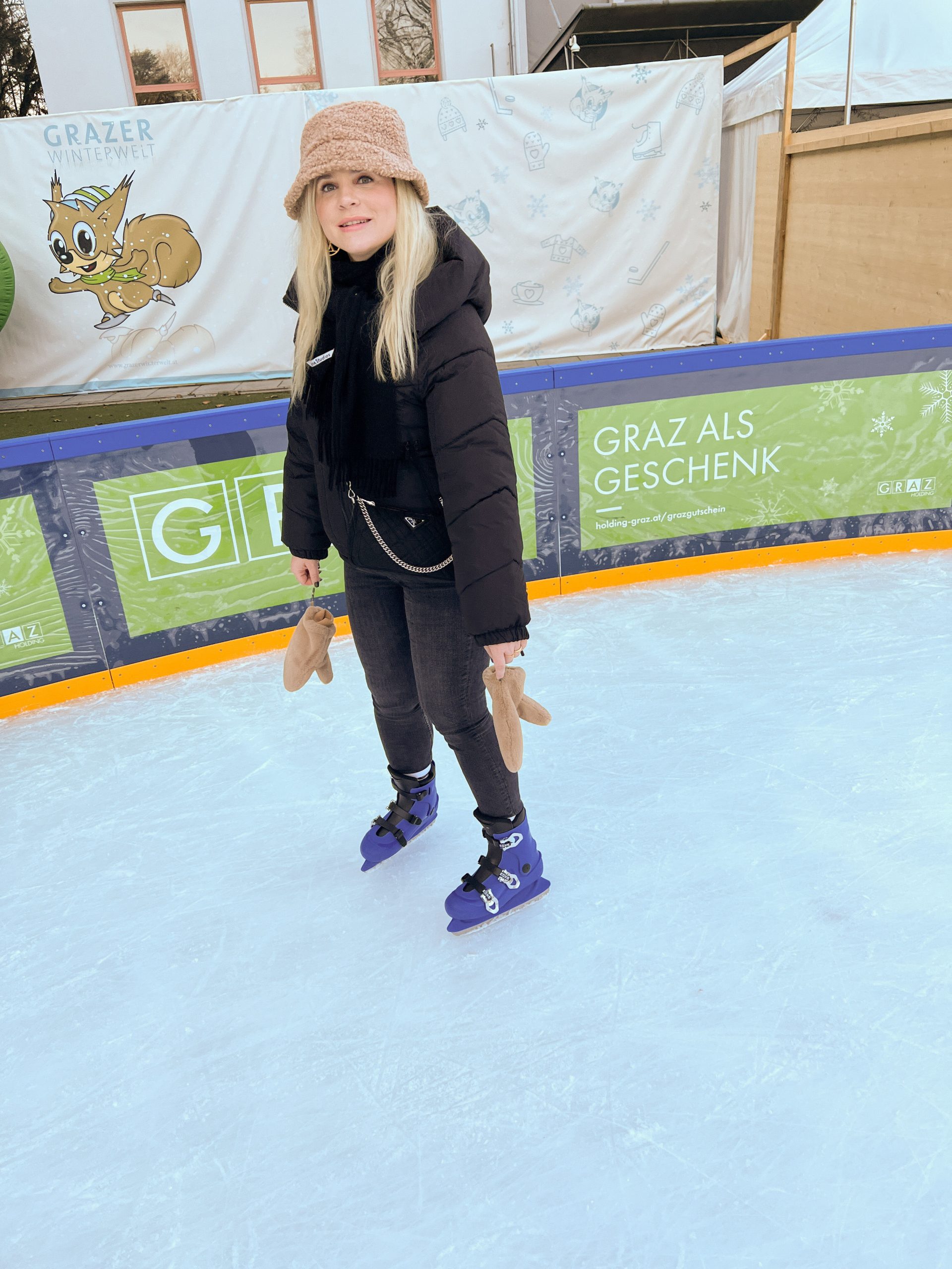 junge Frau mit schwarzer Jacke, schwarzem Schal und beigen Handschuhen und Mütze sowie blauen Eislaufschuhen am Eislaufplatz der Grazer Winterwelt