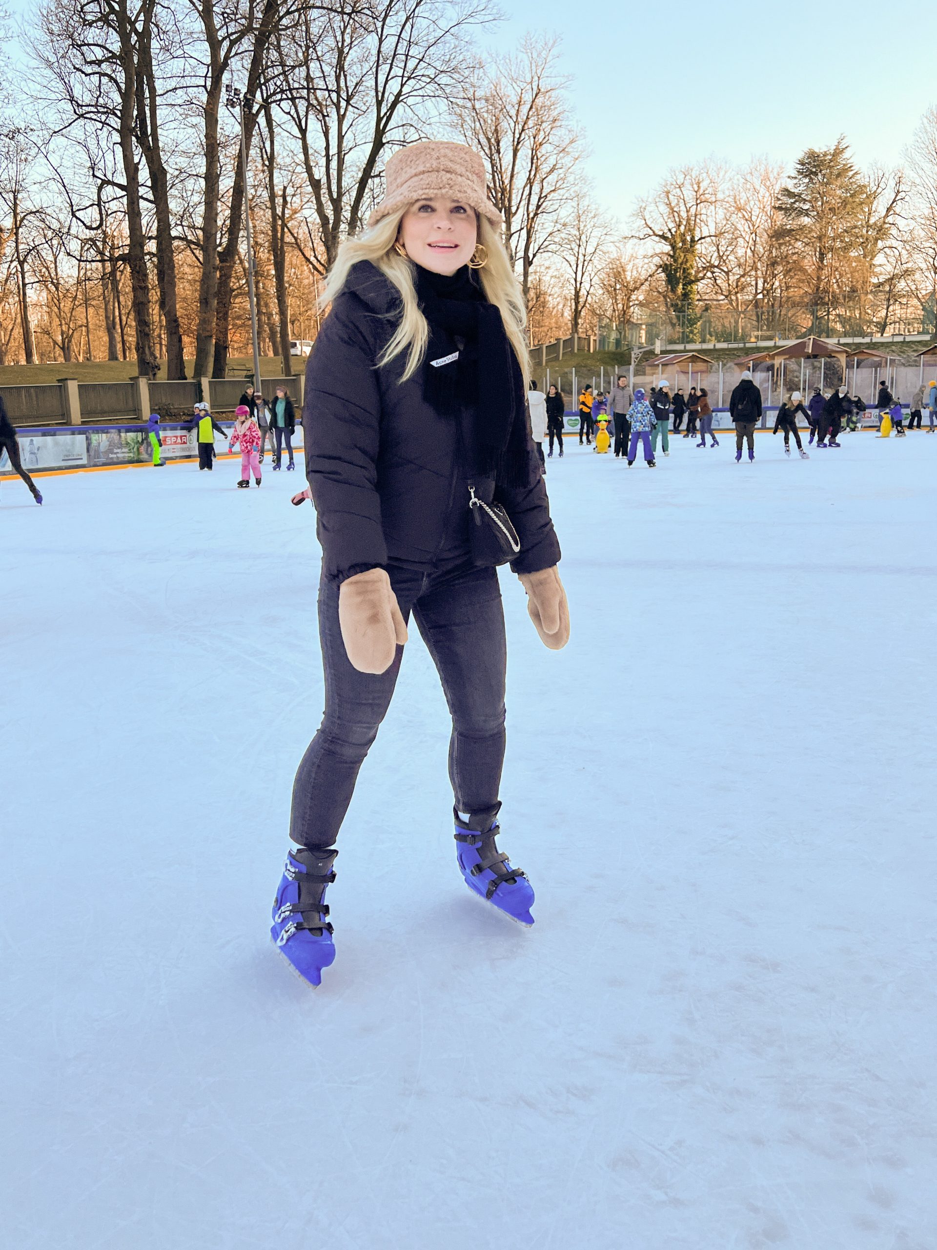 junge Frau mit schwarzer Jacke, schwarzem Schal und beigen Handschuhen und Mütze sowie blauen Eislaufschuhen am Eislaufplatz der Grazer Winterwelt, im Hintergrund andere Eisläuferinnen und Eisläufer