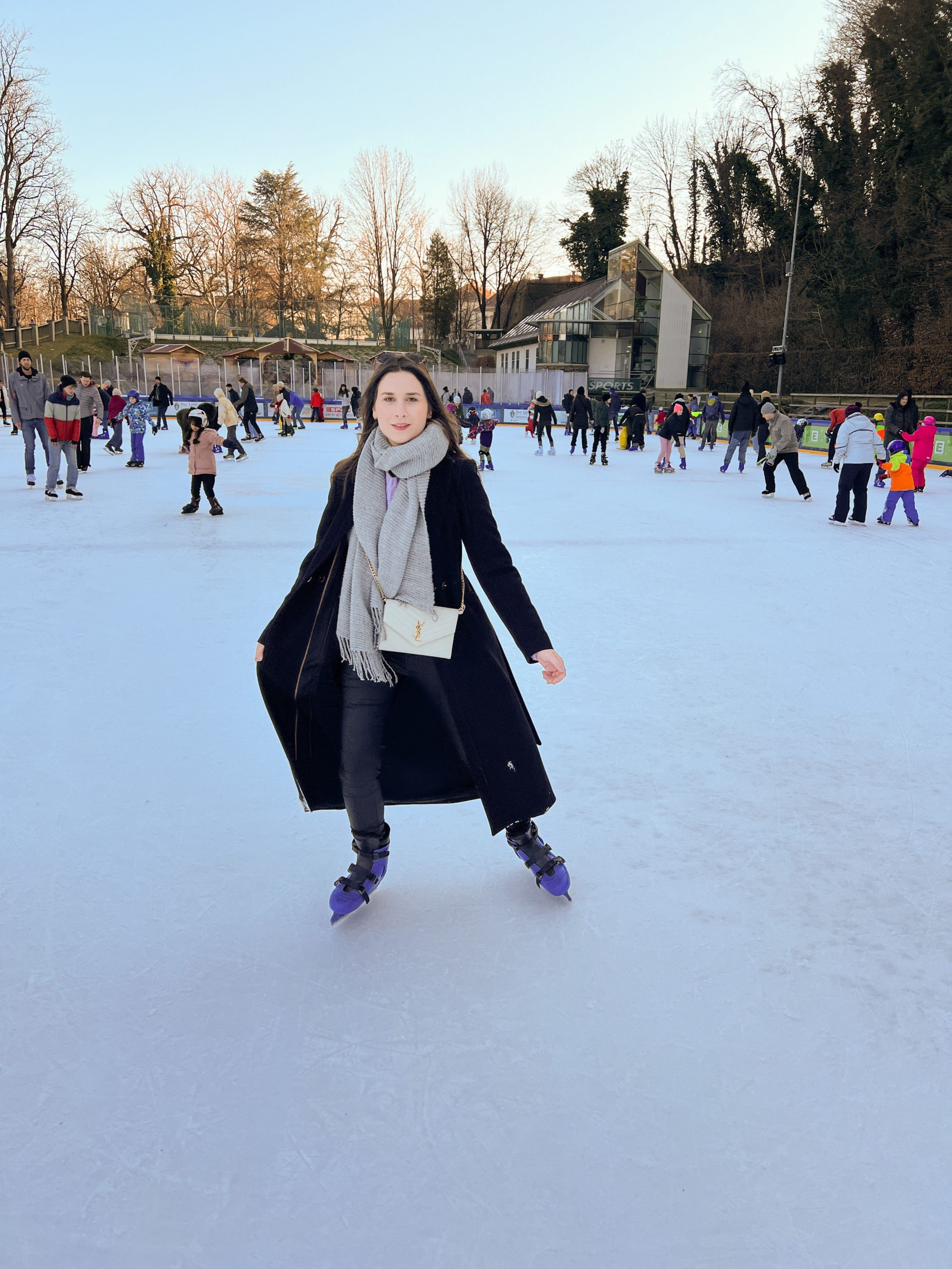 junge Frau mit schwarzem Mantel, grauem Schal und beiger Yves Saint Laurent Tasche und Eislaufschuhen am Eislaufplatz der Grazer Winterwelt, im Hintergrund andere Eisläuferinnen und Eisläufer