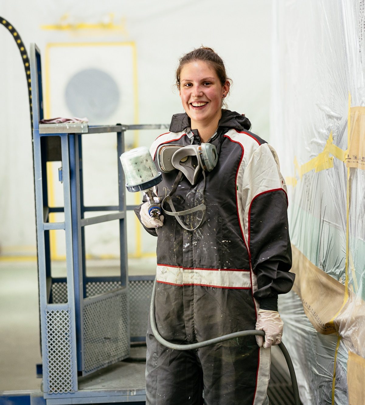 Lächelnde Frau in Schutzkleidung hält Lackierpistole in Werkstatt für Fahrzeuglackierungen.