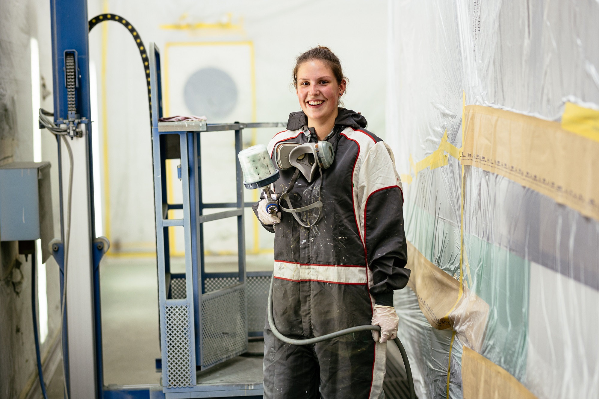 Lächelnde Frau in Schutzkleidung hält Lackierpistole in Werkstatt für Fahrzeuglackierungen.