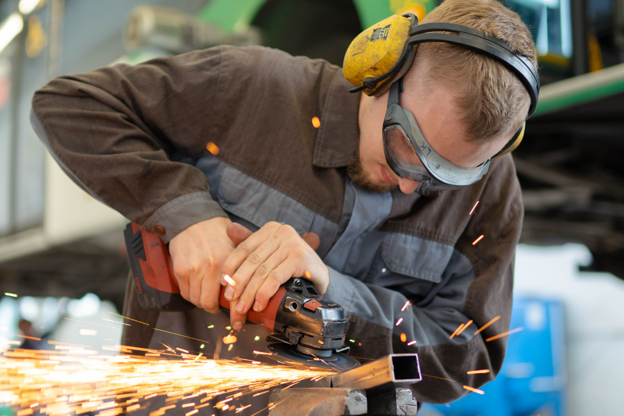 Junger Mann mit Schutzbrille und Gehörschutz arbeitet mit Schleifmaschine an Metallstück.
