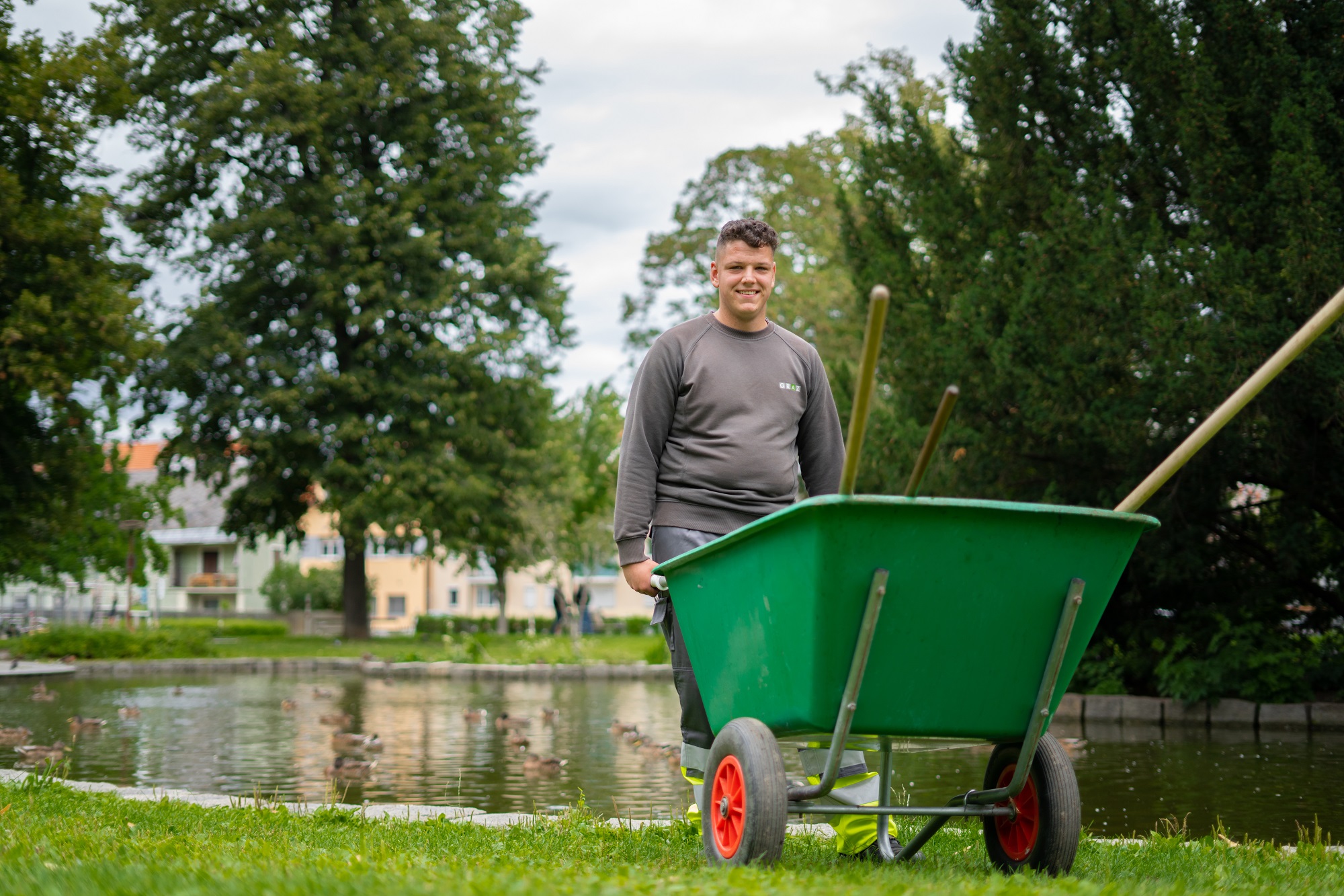Junger Mann mit grüner Schubkarre in einem Park vor Teich mit Bäumen im Hintergrund