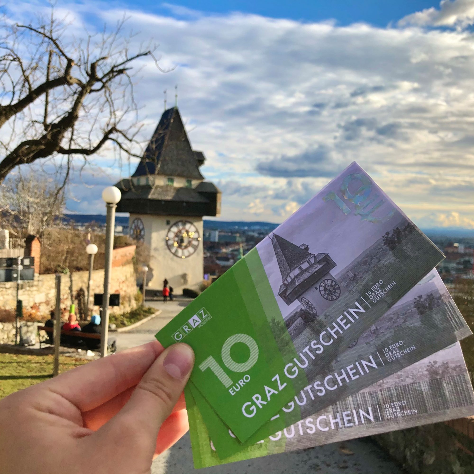 A hand holds three "Graz Gutschein" vouchers, each worth 10 Euro, in front of the Graz Clock Tower.