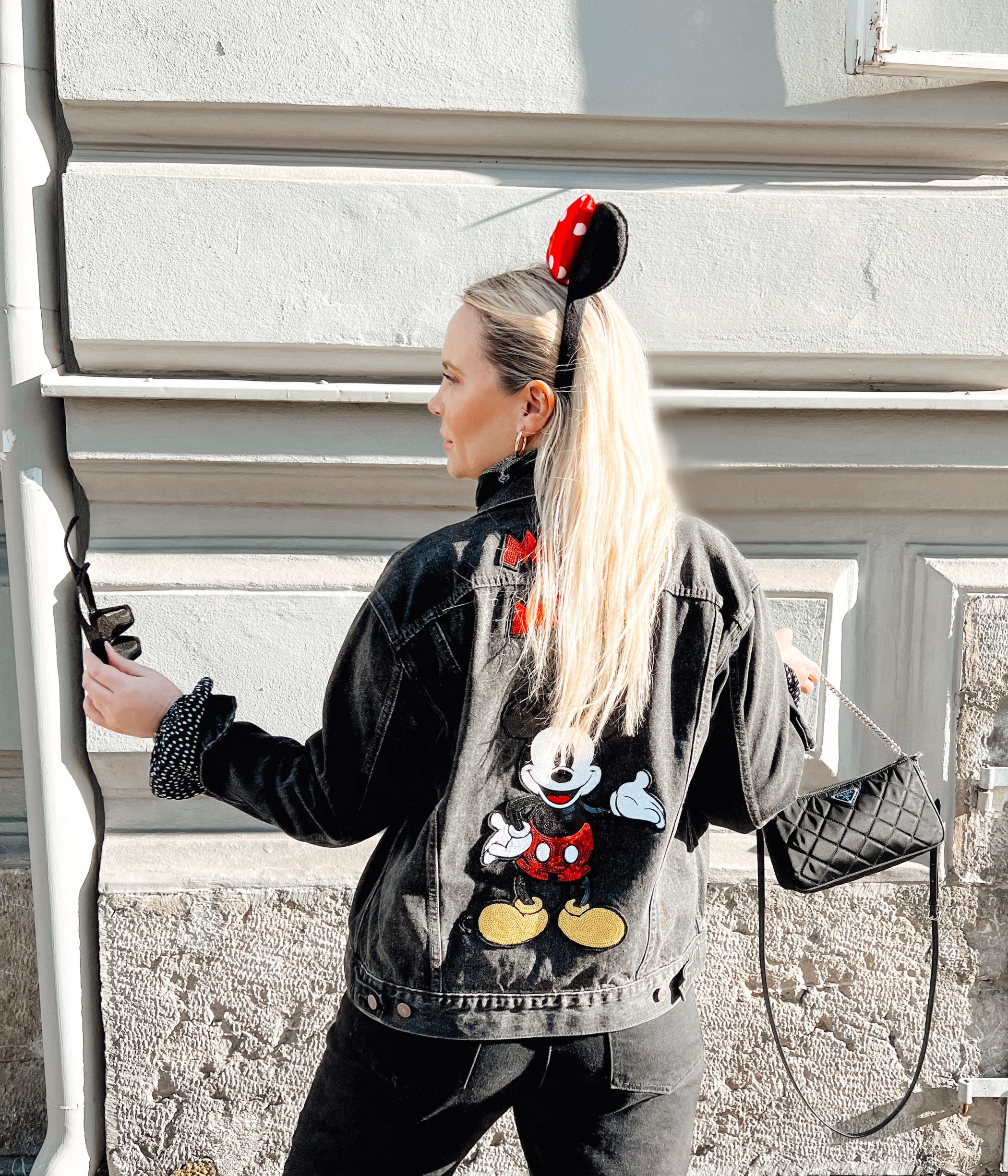 junge blonde Frau vor einer Hausmauer mit Mickey Mouse Ohren als Haarreifen und einer Jeansjacke mit Mickey Mouse Abbildung am Rücken