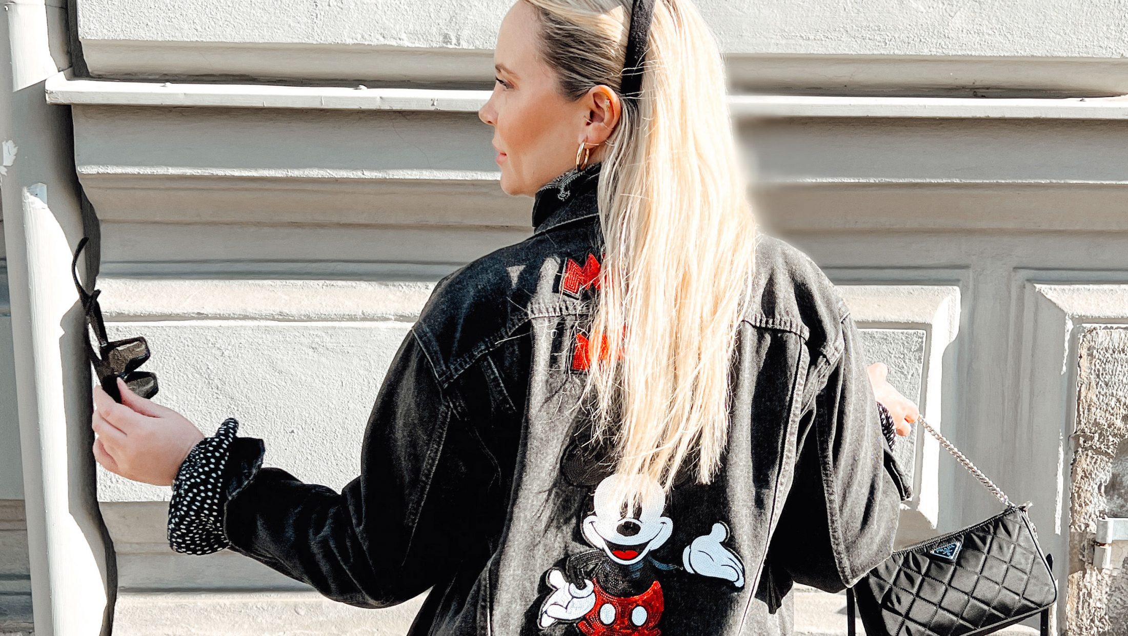 junge blonde Frau vor einer Hausmauer mit Mickey Mouse Ohren als Haarreifen und einer Jeansjacke mit Mickey Mouse Abbildung am Rücken