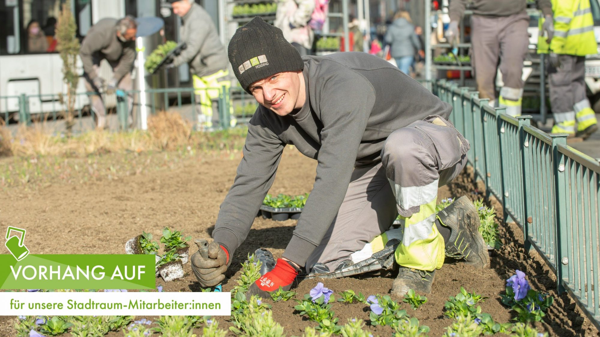 Gärtner pflanzt Blumen in Beet und lächelt in die Kamera, im Hintergrund Kollegen bei der Arbeit.