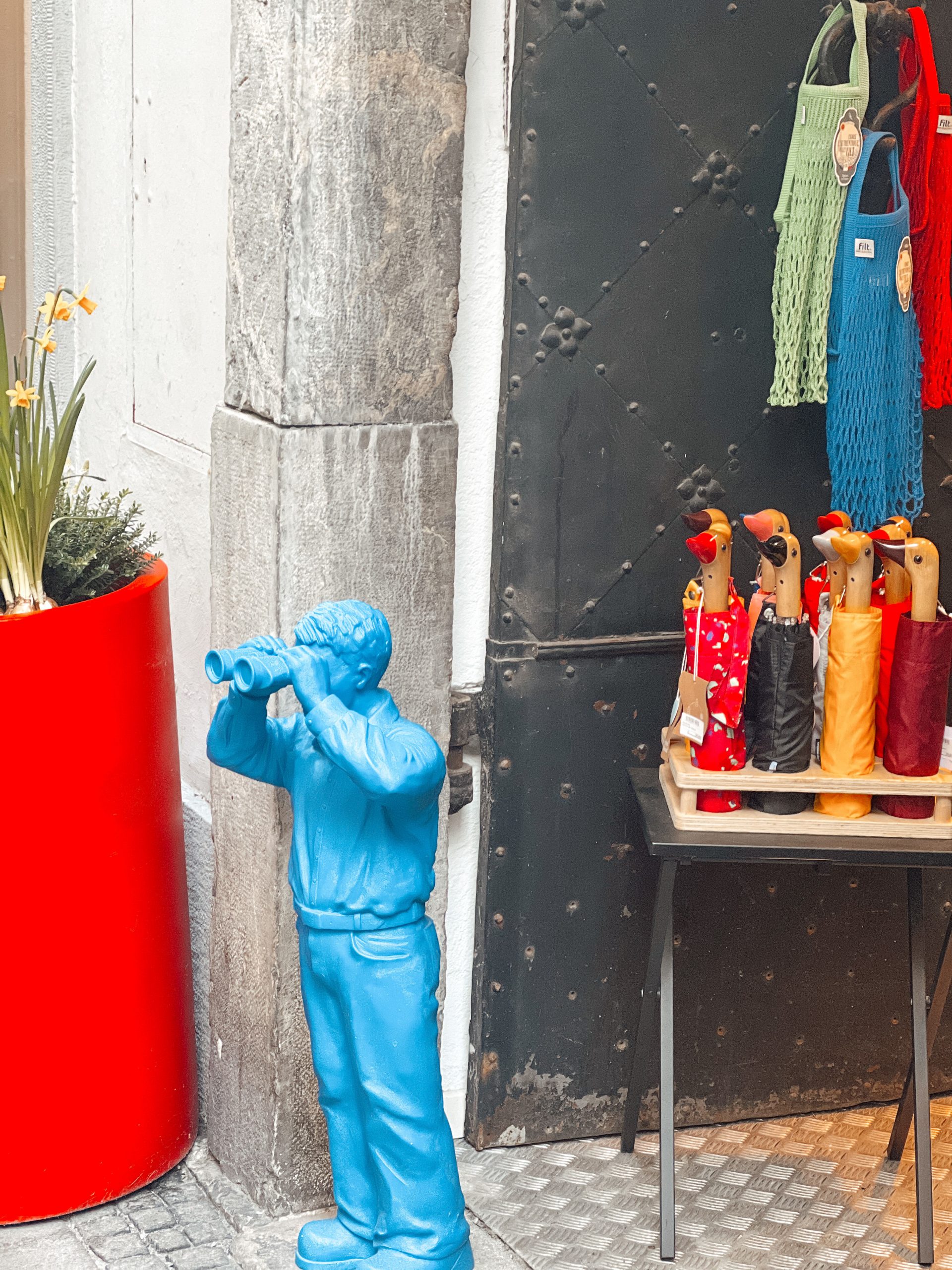 blaue kleine Figur eines Mannes, der durch ein Fernglas schaut steht neben einem Tischchen voll bunter Enten-Regenschirme vor dem MuR - Design Store