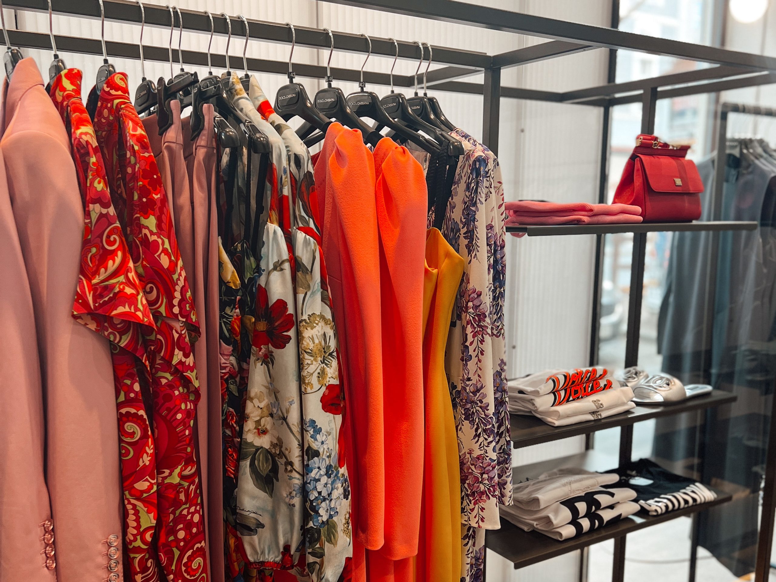 Kleiderstange mit verschiedenen bunten Kleidern, Blusen und Blazern (einfarbig und mit floralen Prints) neben einem Regal mit T-Shirts, Sandalen und einer roten Tasche im Geschäft KNILLI