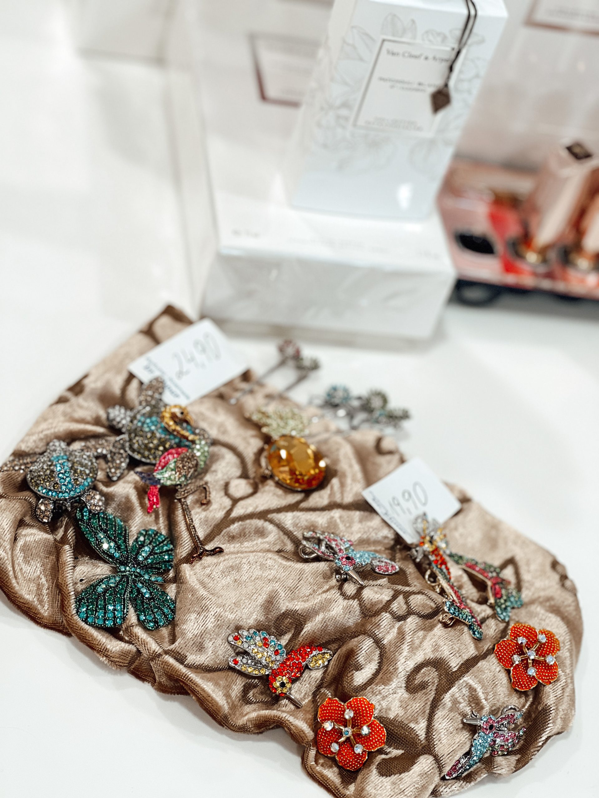verschiedene Broschen und Haarklammern mit Schmetterlingen, Blumen und Vögeln auf einem Tuch, das auf einer weißen Fläche liegt, im Hintergrund Parfums und Lippenstifte bei Parfümerie Dr. Ebner