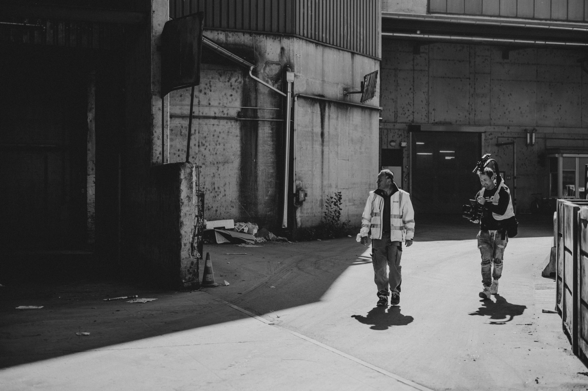 Schwarz-Weiß-Foto: Arbeiter in Sicherheitsjacke und Kameramann in einer Industrieanlage.