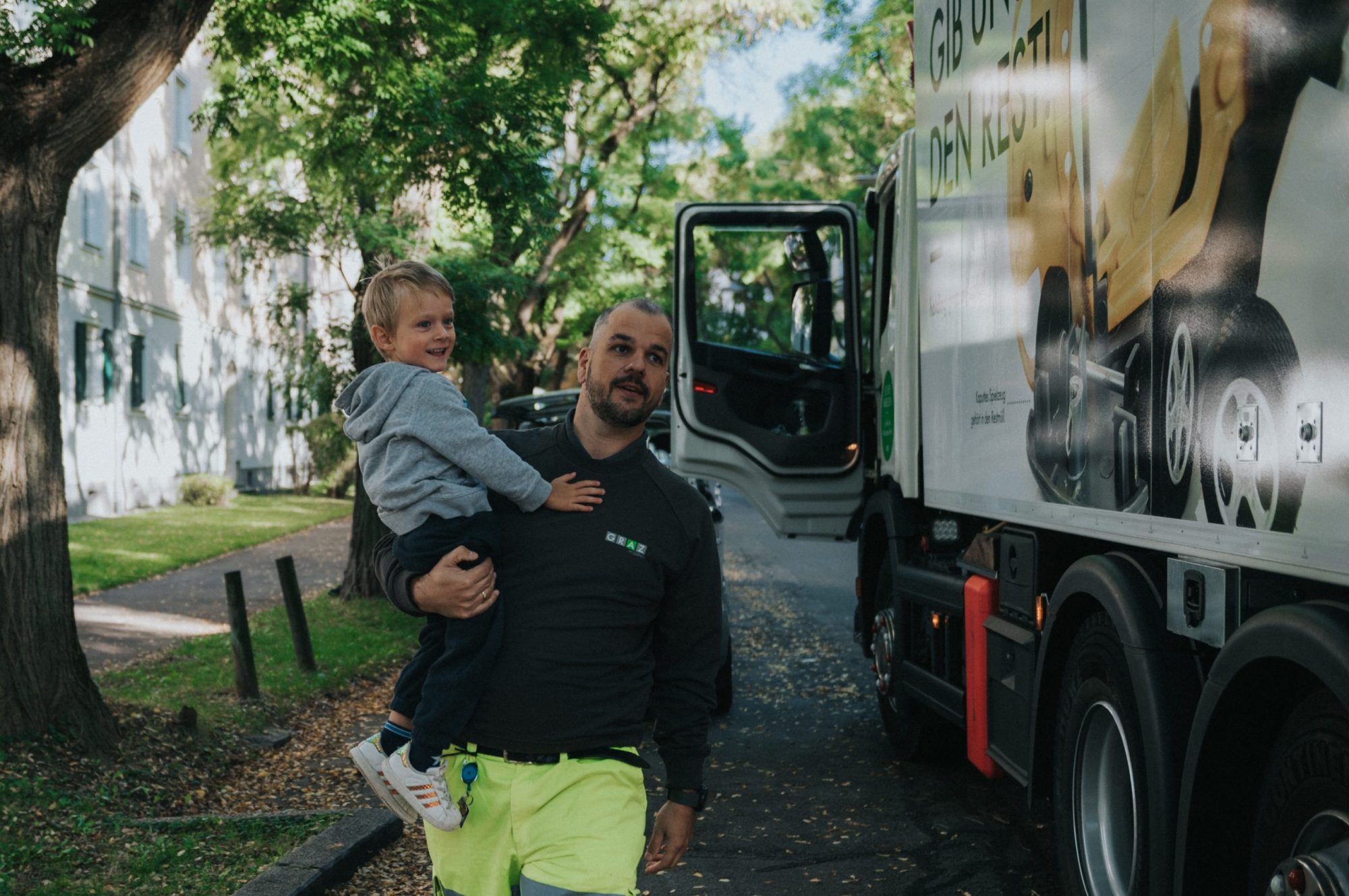 Mitarbeiter der Abfallwirtschaft mit kleinem Buben am Arm geht entlang eines Müllfahrzeuges