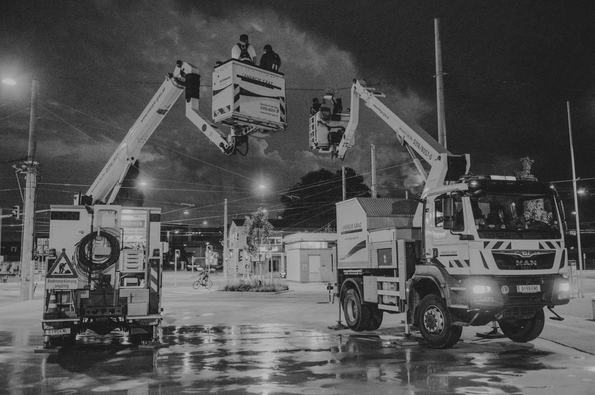 Schwarz-Weiß-Foto: Arbeitsfahrzeuge mit Hebebühnen, Arbeiter reparieren Oberleitungen bei Nacht.