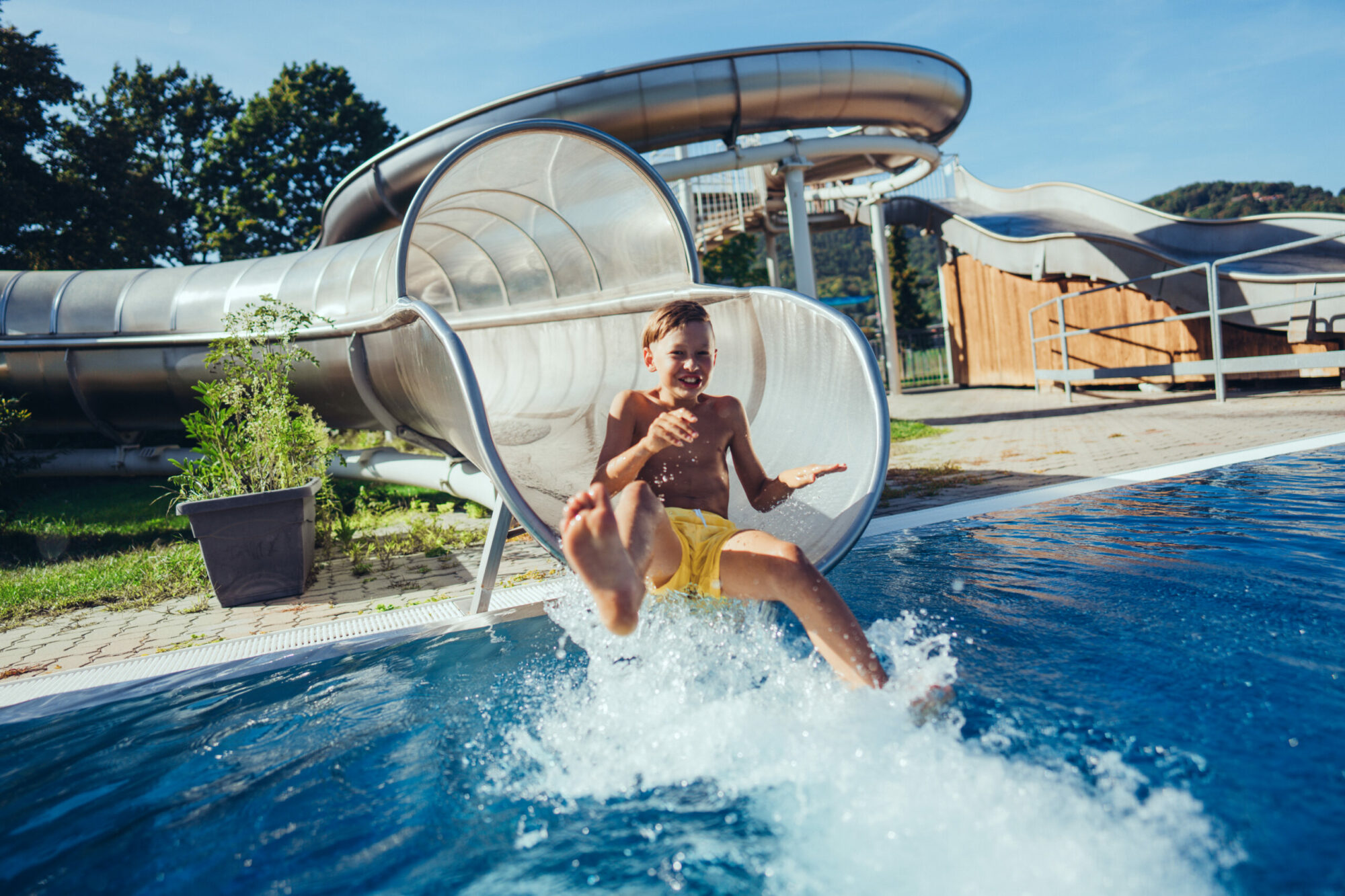 Bub rutscht lachend bei sonnigem Wetter aus einer Wasserrutsche in ein Becken.