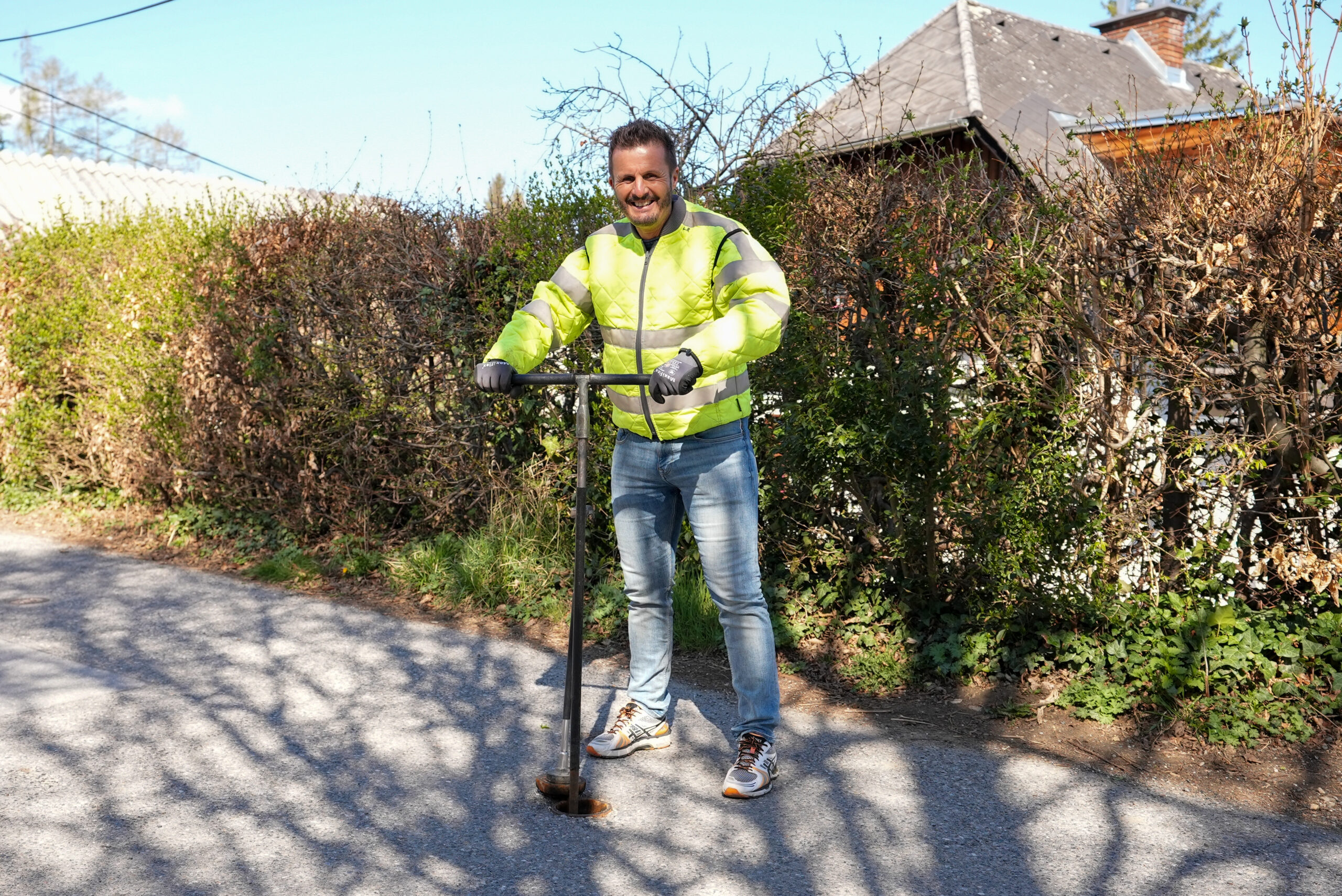 Mann in Warnjacke steht lächelnd mit Werkzeug auf Straße vor grüner Hecke und blauem Himmel.