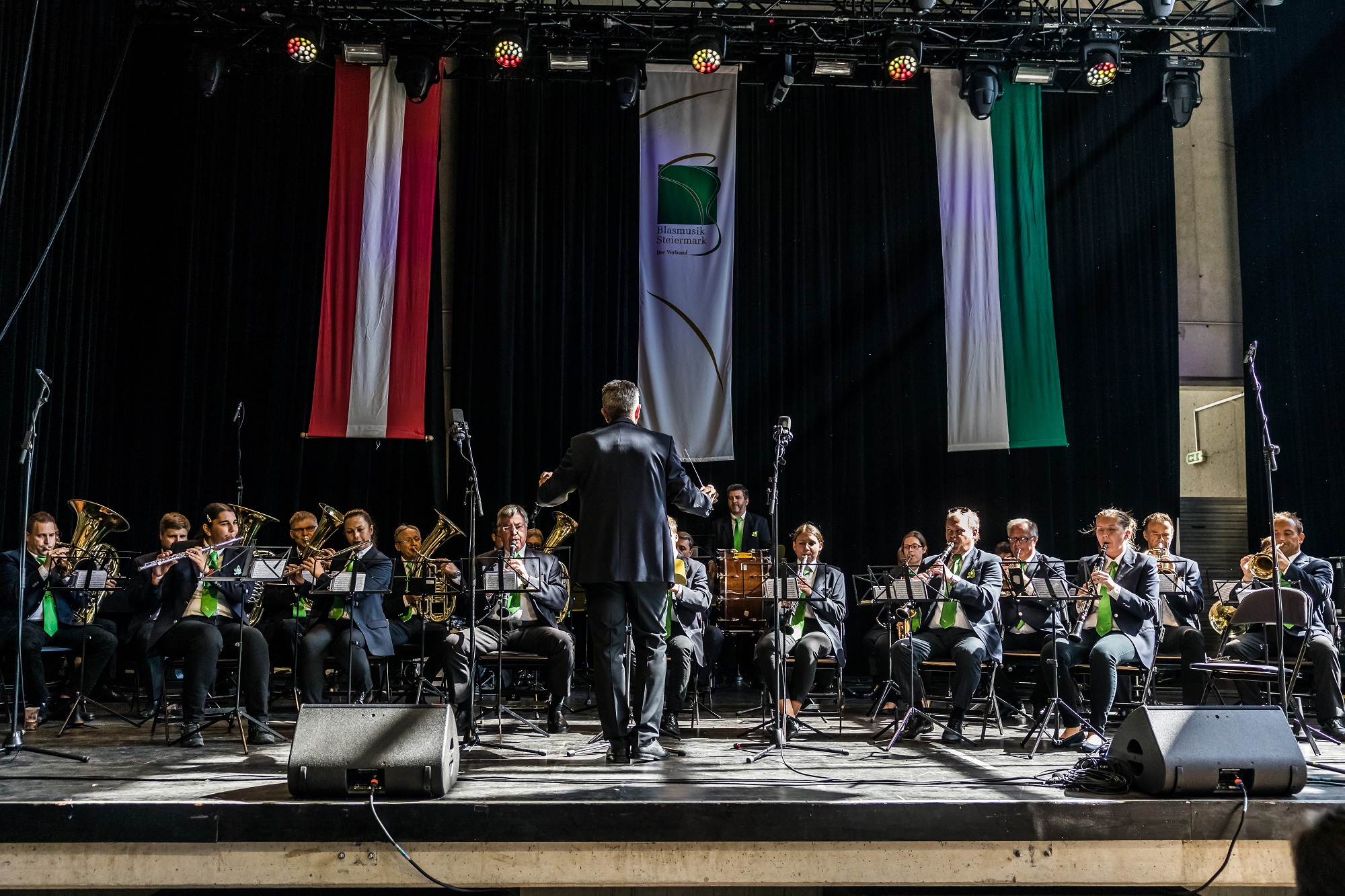 Blasorchester spielt auf Bühne, dirigiert von einem Mann, vor rot-weißen und grünen Fahnen.