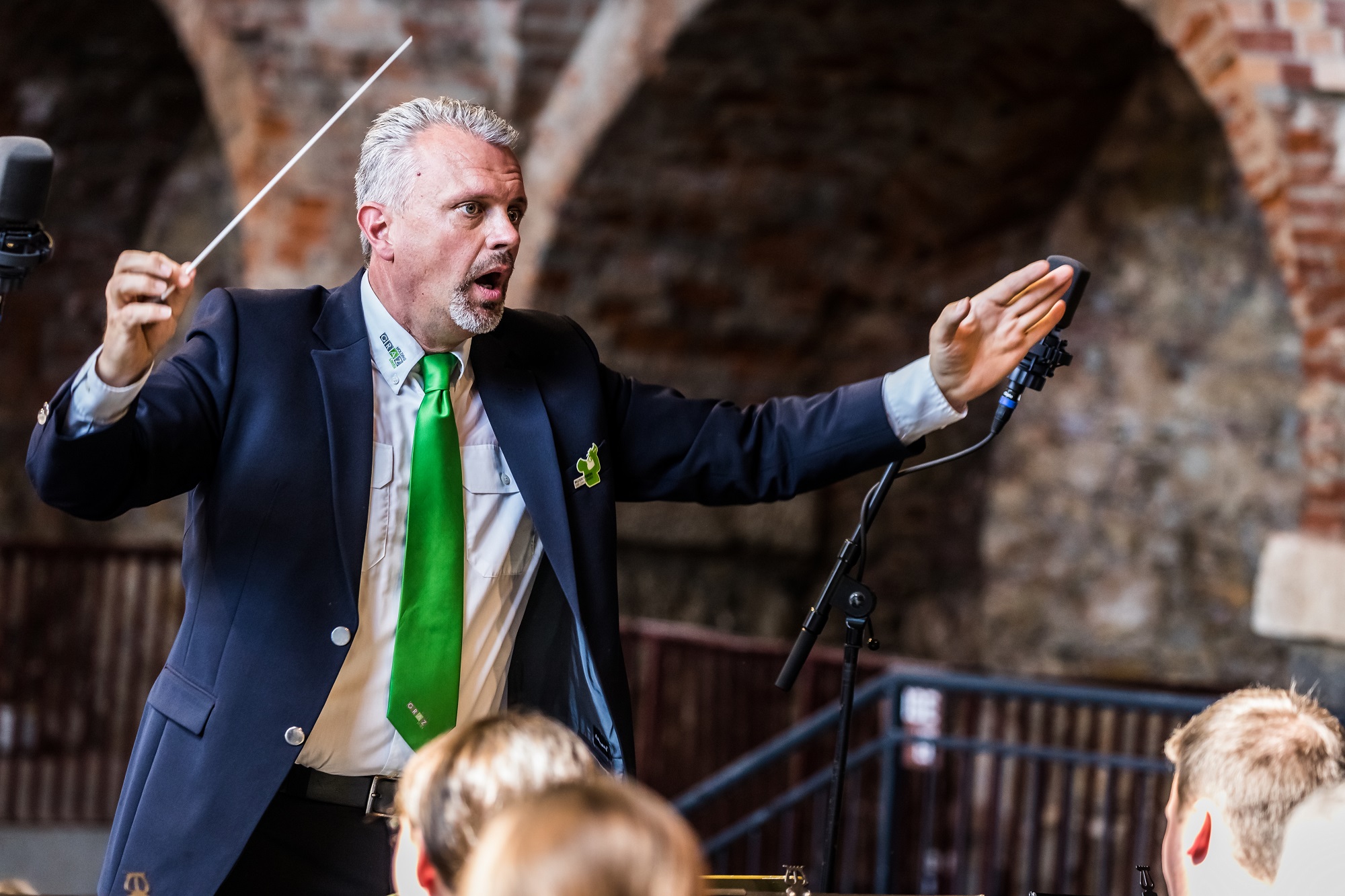 Dirigent mit grüner Krawatte leitet Orchester vor historischer Backsteinwand.