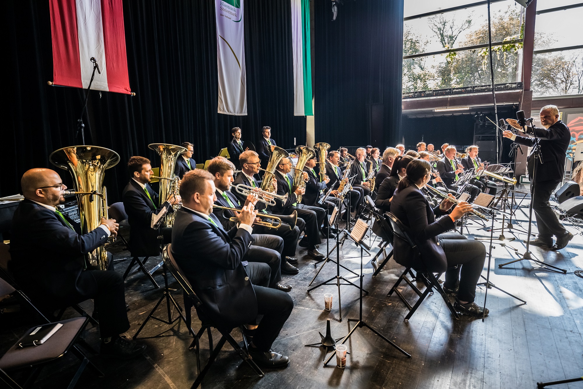 Orchester spielt auf Bühne mit Tubas und Trompeten, dirigiert von Mann, vor großen Fenstern.