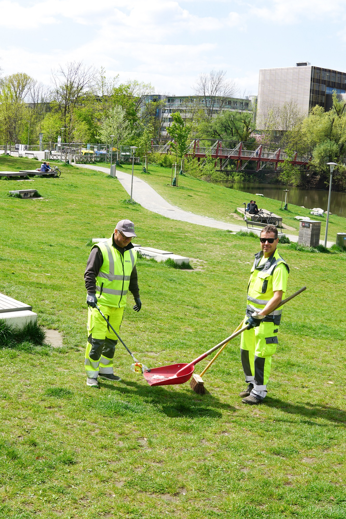 Zwei Arbeiter in Warnwesten reinigen eine Grünfläche in einem Park bei sonnigem Wetter.