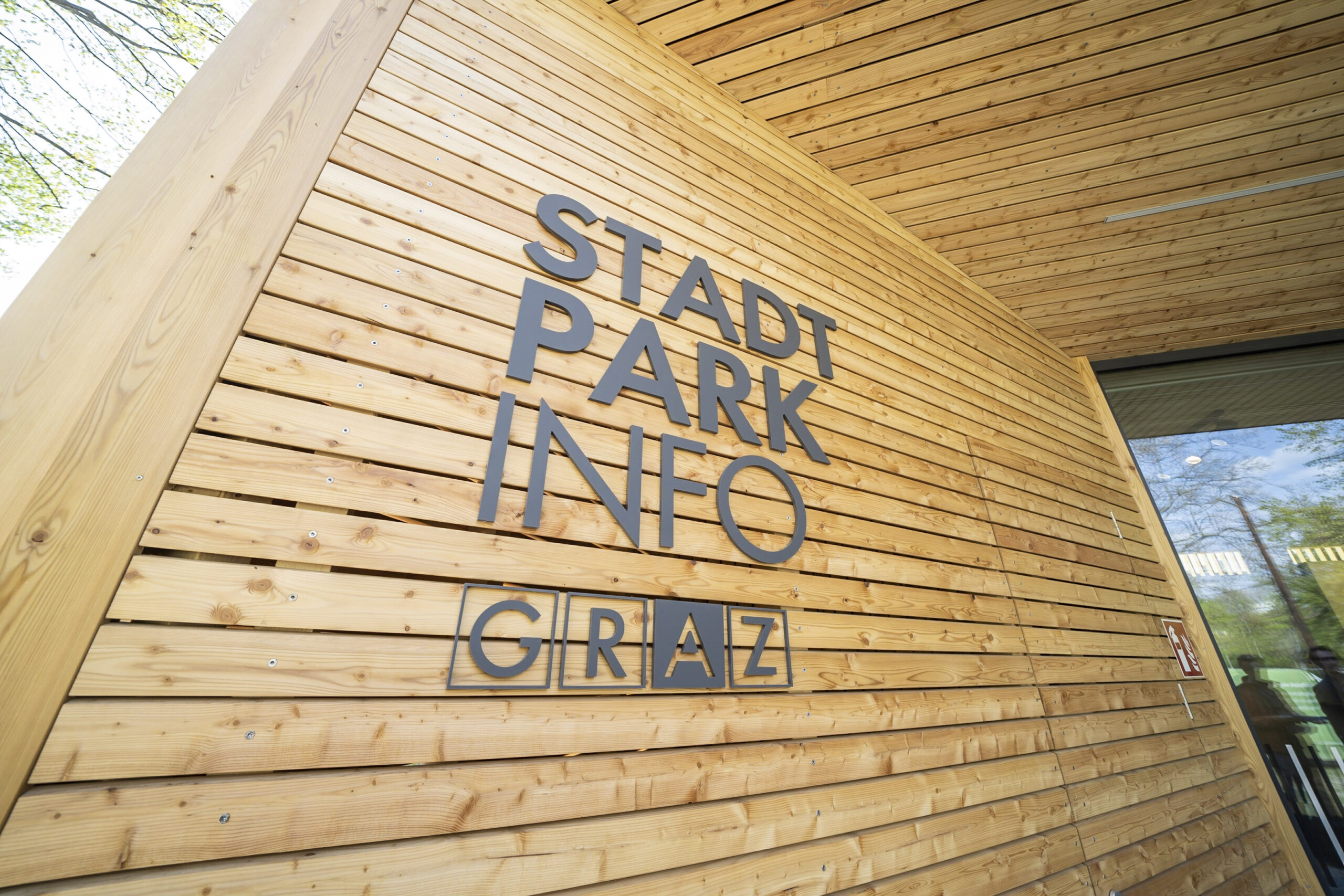 Holzgebäude mit der Aufschrift "Stadtpark Info Graz" und moderner Architektur.