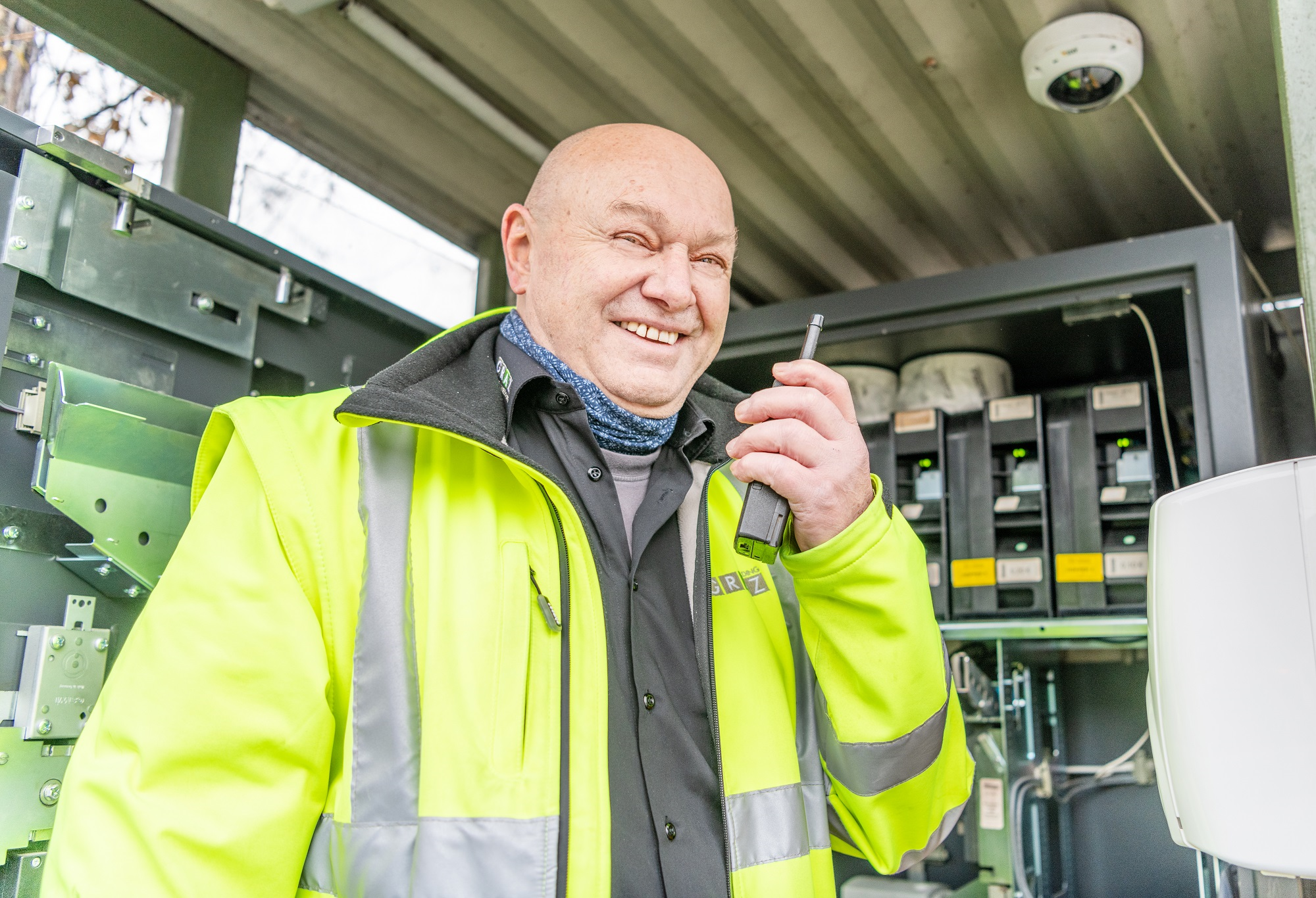 Mann in Warnweste spricht lächelnd in Funkgerät, im Hintergrund technische Anlagen.