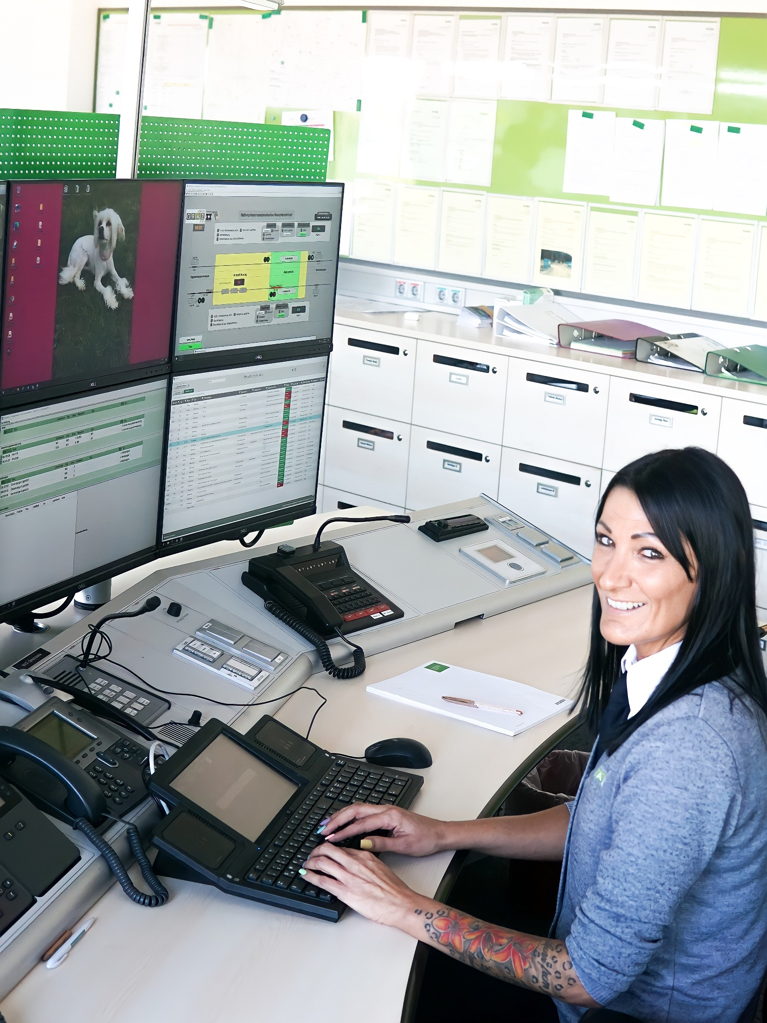 Frau in Leitstelle mit mehreren Monitoren und Telefonen, lächelt in die Kamera.