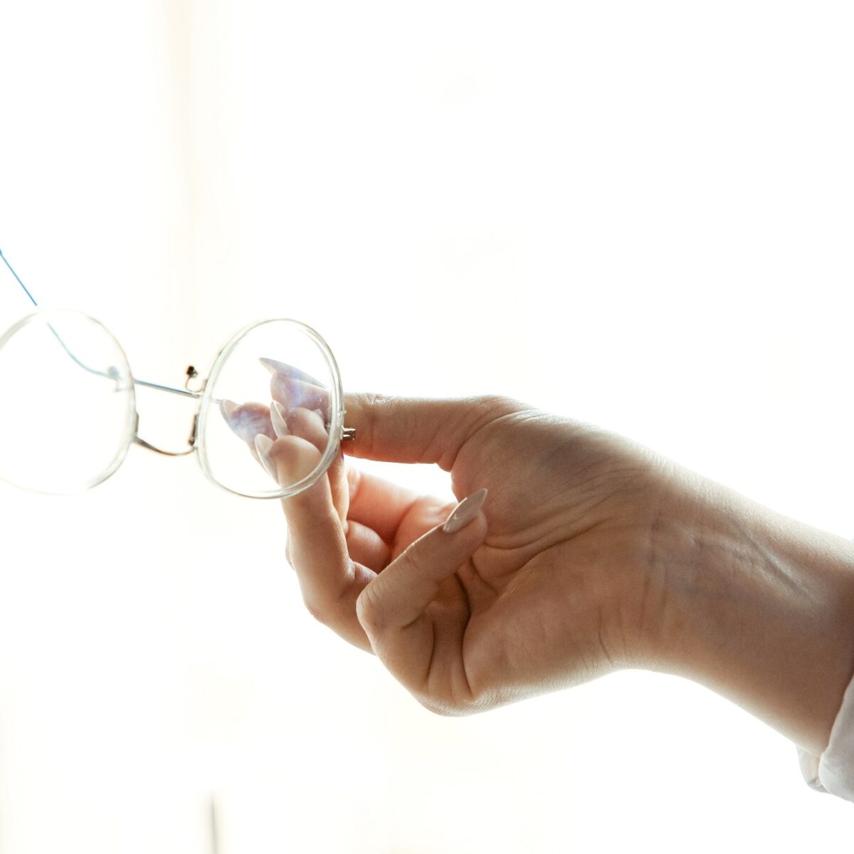 Hand hält eine runde Brille vor einem hellen Hintergrund, feine Details der Gläser sichtbar.