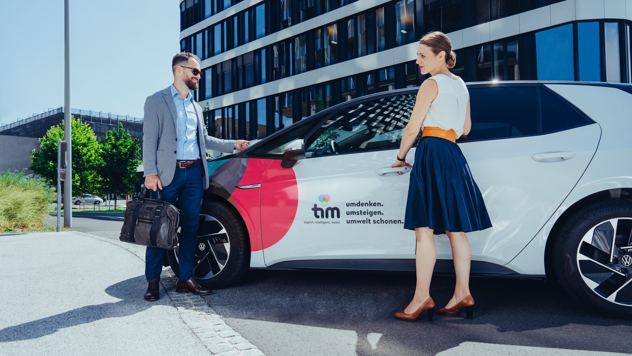 Mann und Frau stehen vor einem tim-Carsharing-Auto vor modernem Bürogebäude.