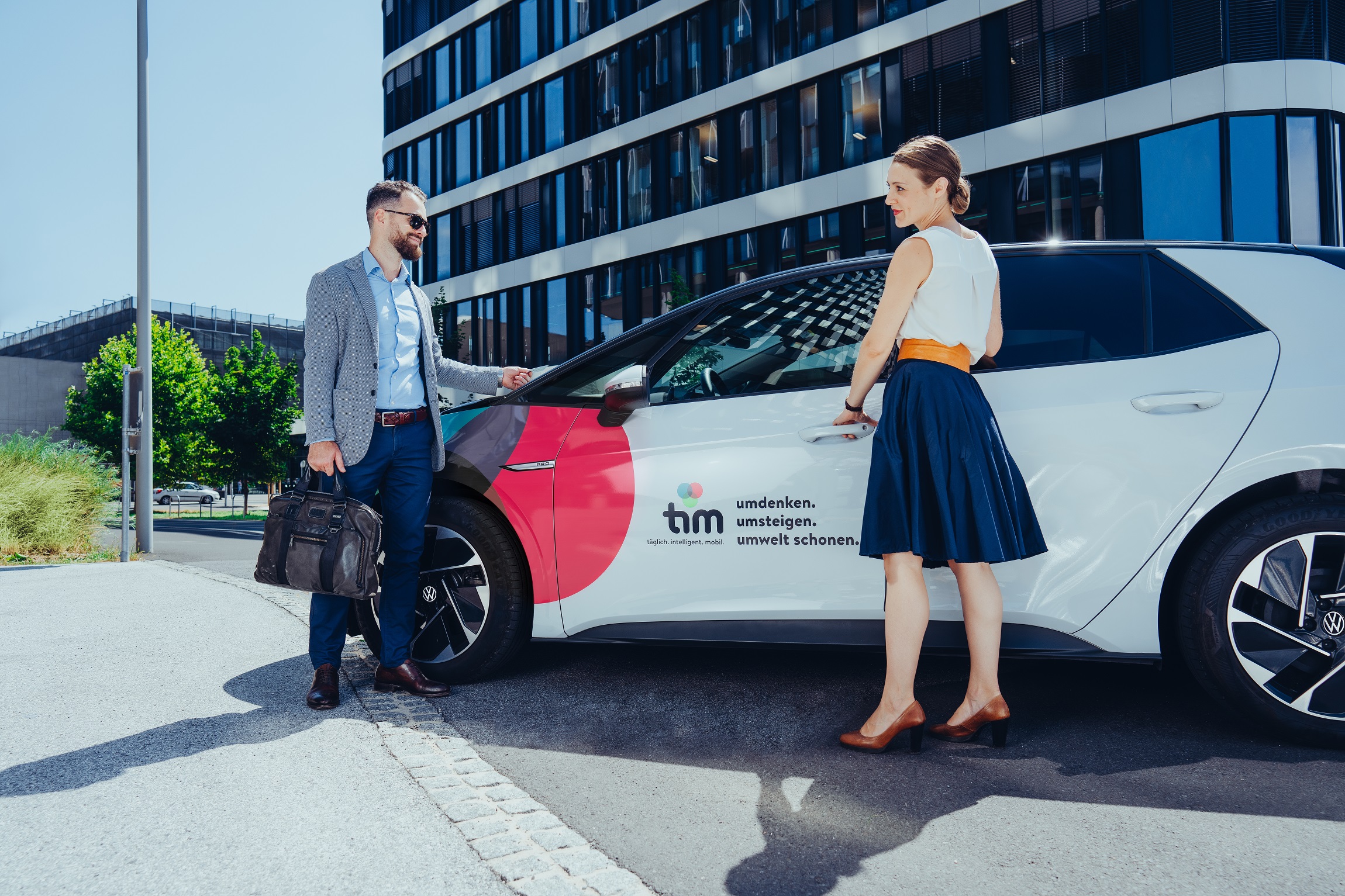 Mann und Frau stehen vor einem tim-Carsharing-Auto vor modernem Bürogebäude.