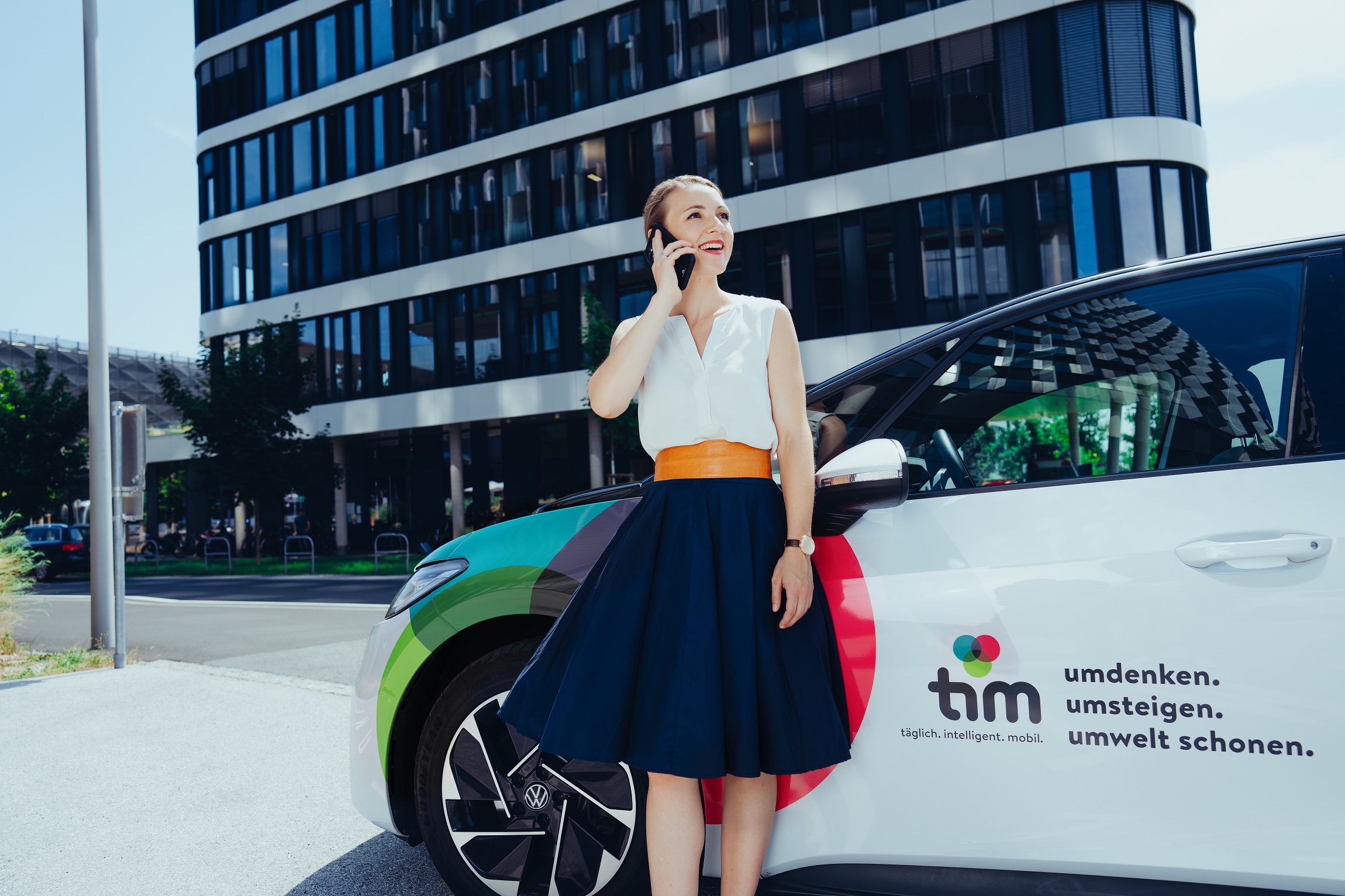 Frau telefoniert lächelnd neben tim-Carsharing-Auto vor modernem Bürogebäude.