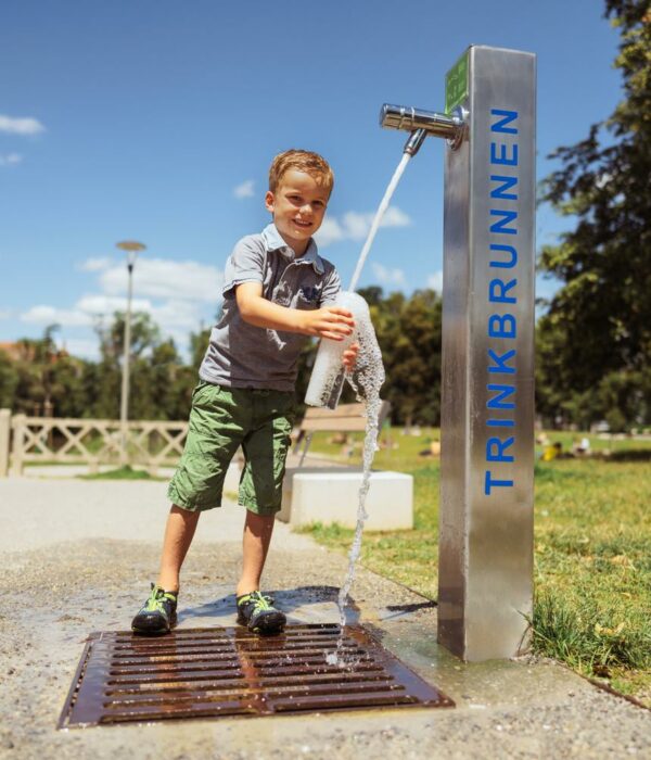 Buf füllt eine Flasche an einem Trinkbrunnen in einem Park bei sonnigem Wetter.