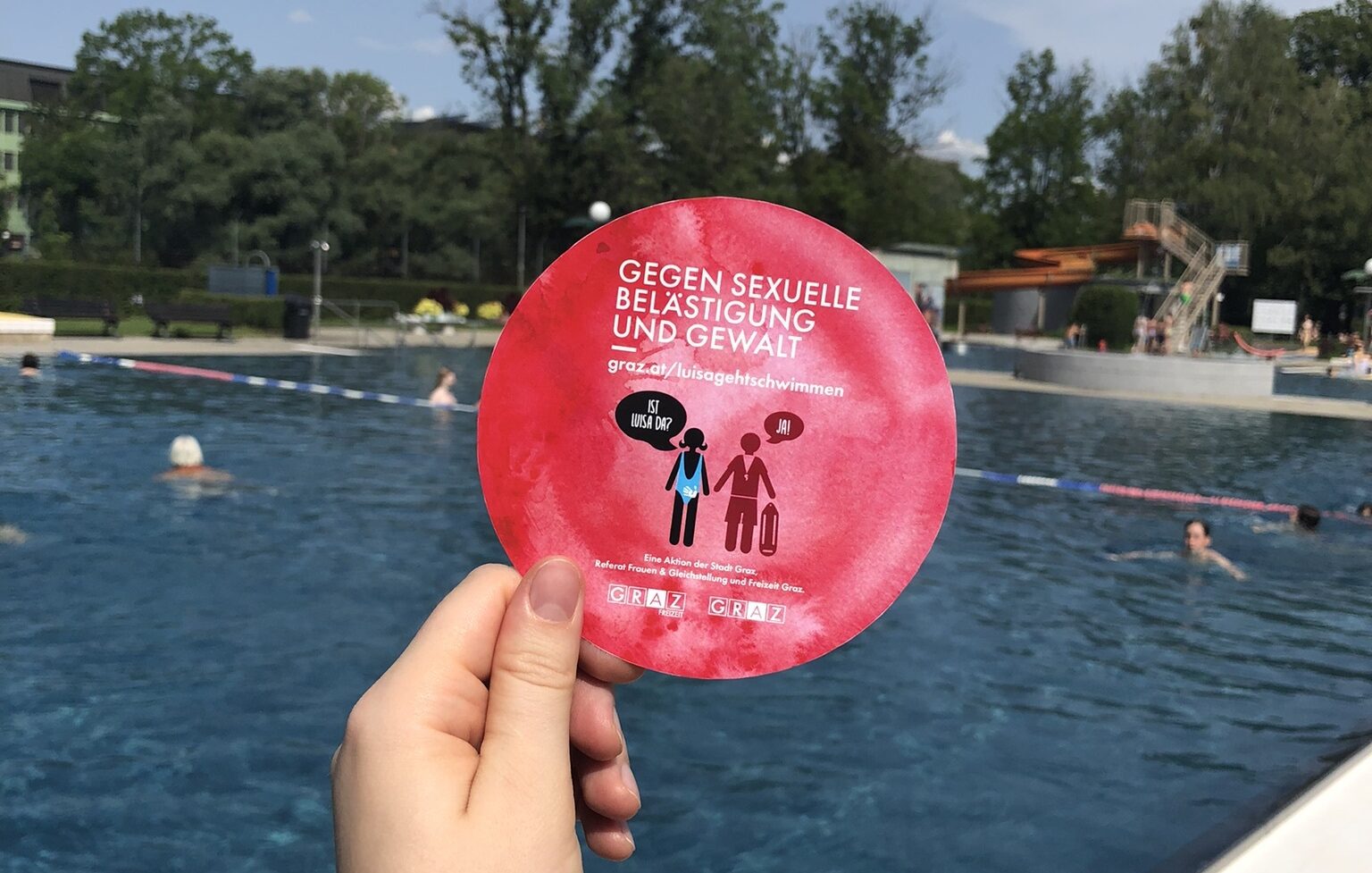 Hand hält roten Flyer mit Botschaft gegen sexuelle Belästigung, dahinter Freibad mit Schwimmern
