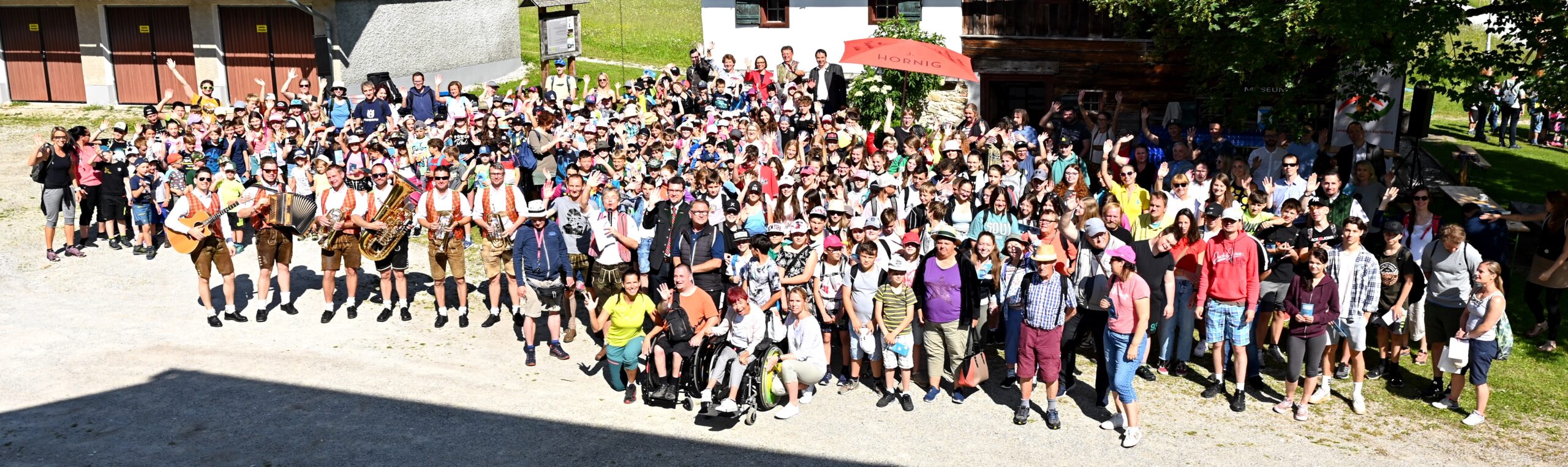 Große Gruppe bei Outdoor-Veranstaltung, im Vordergrund Musikanten in Tracht mit Instrumenten.