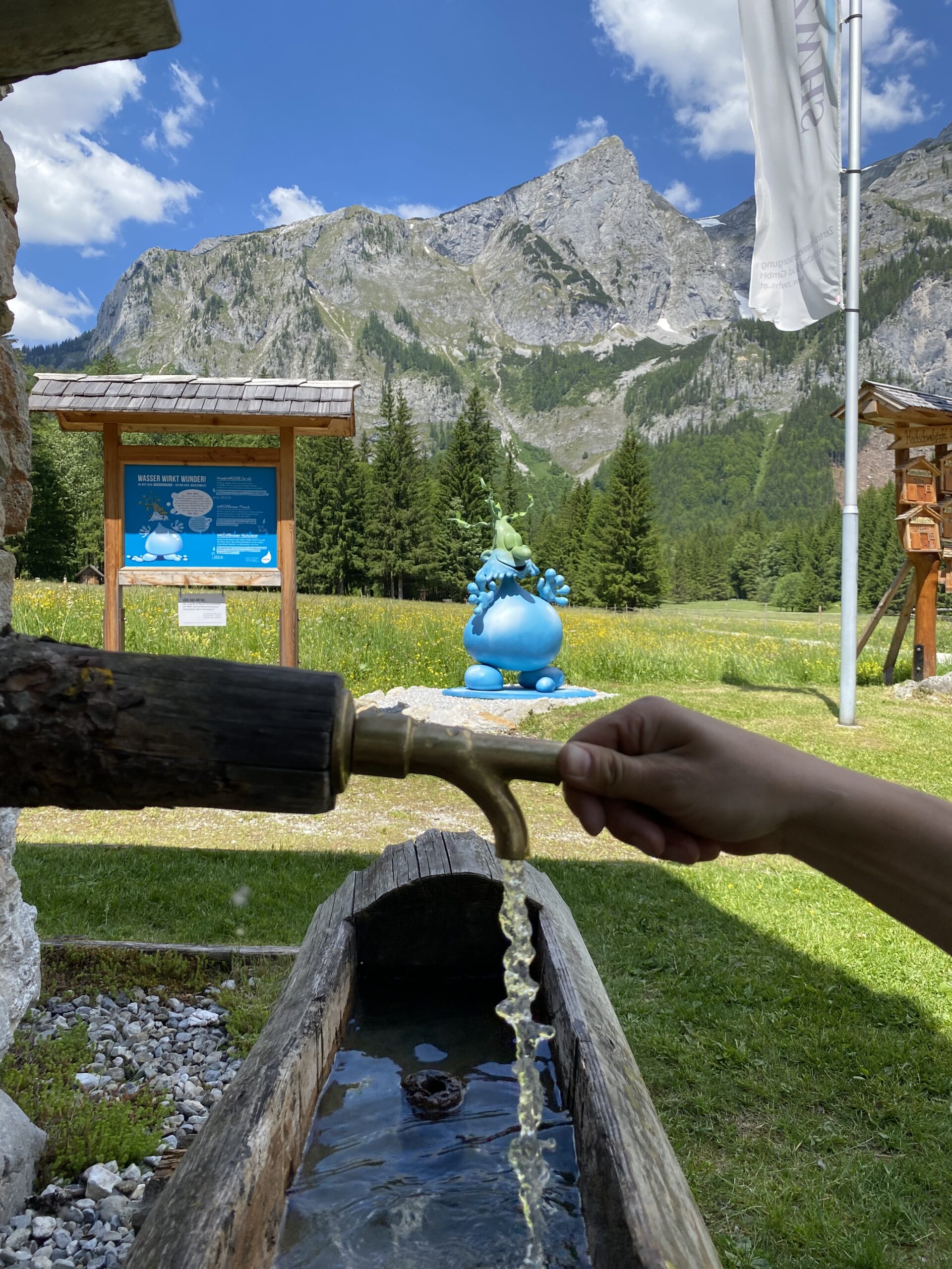 Wasser fließt aus einem Holzbrunnen, im Hintergrund Berge, Wiesen und eine blaue Figur.