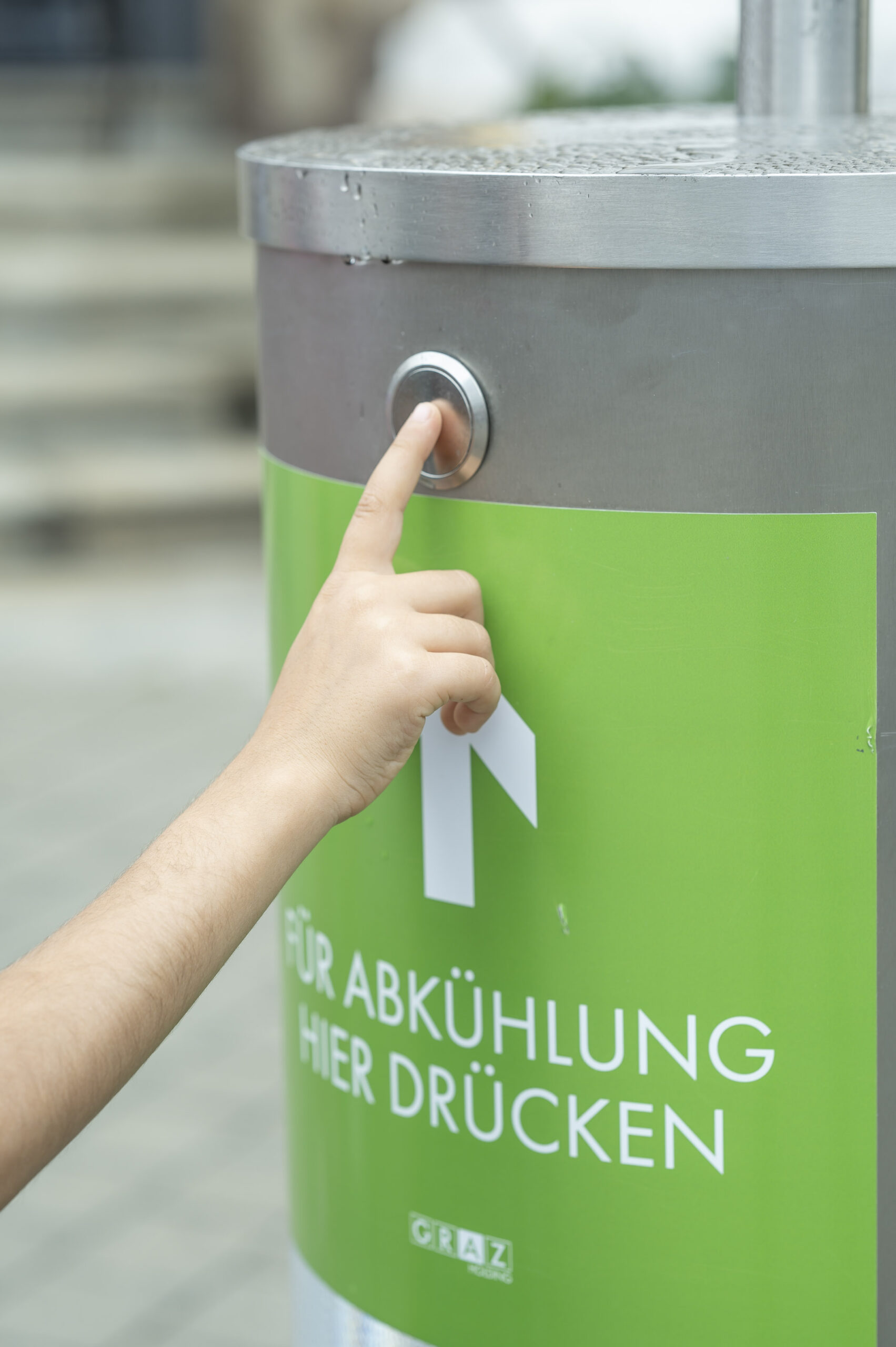 Hand drückt auf Knopf an Abkühlungsstation mit grünem Schild: 'Für Abkühlung hier drücken'.