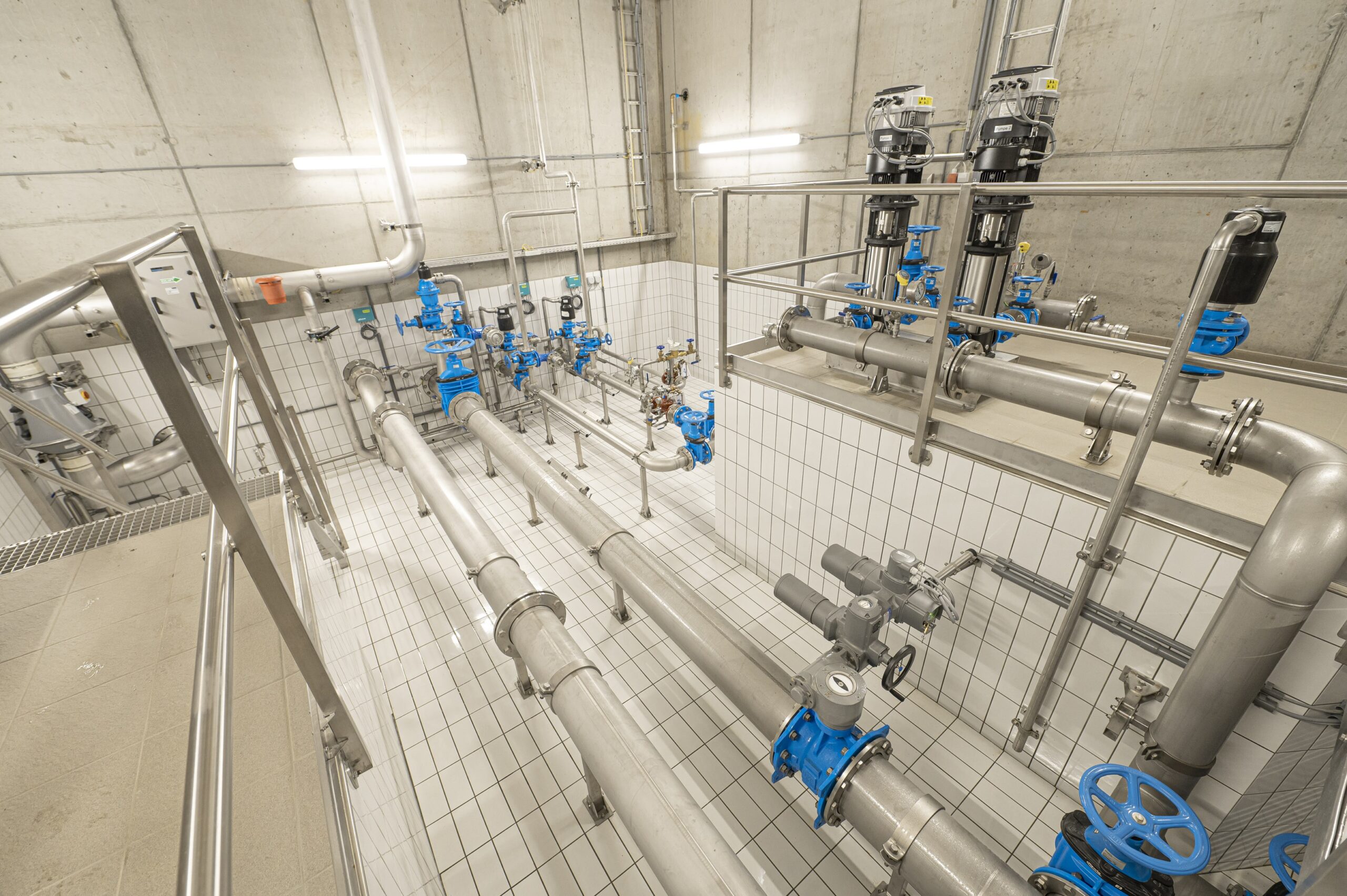 Technische Anlage mit Edelstahlrohren, blauen Ventilen und Fliesenboden in einem hellen Raum.