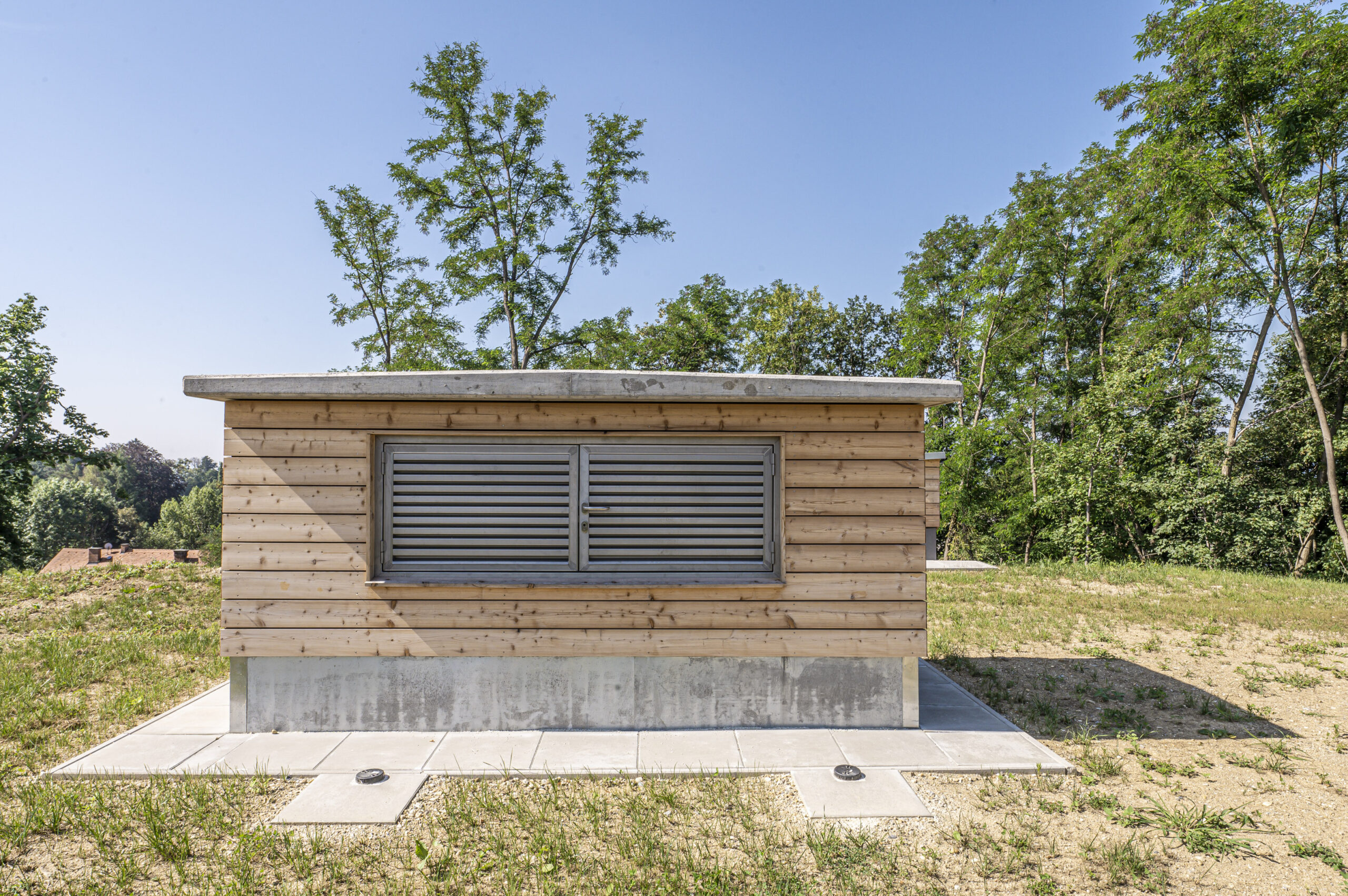 Holzgebäude mit Lüftungsschlitzen, umgeben von Grünflächen und Bäumen unter blauem Himmel.