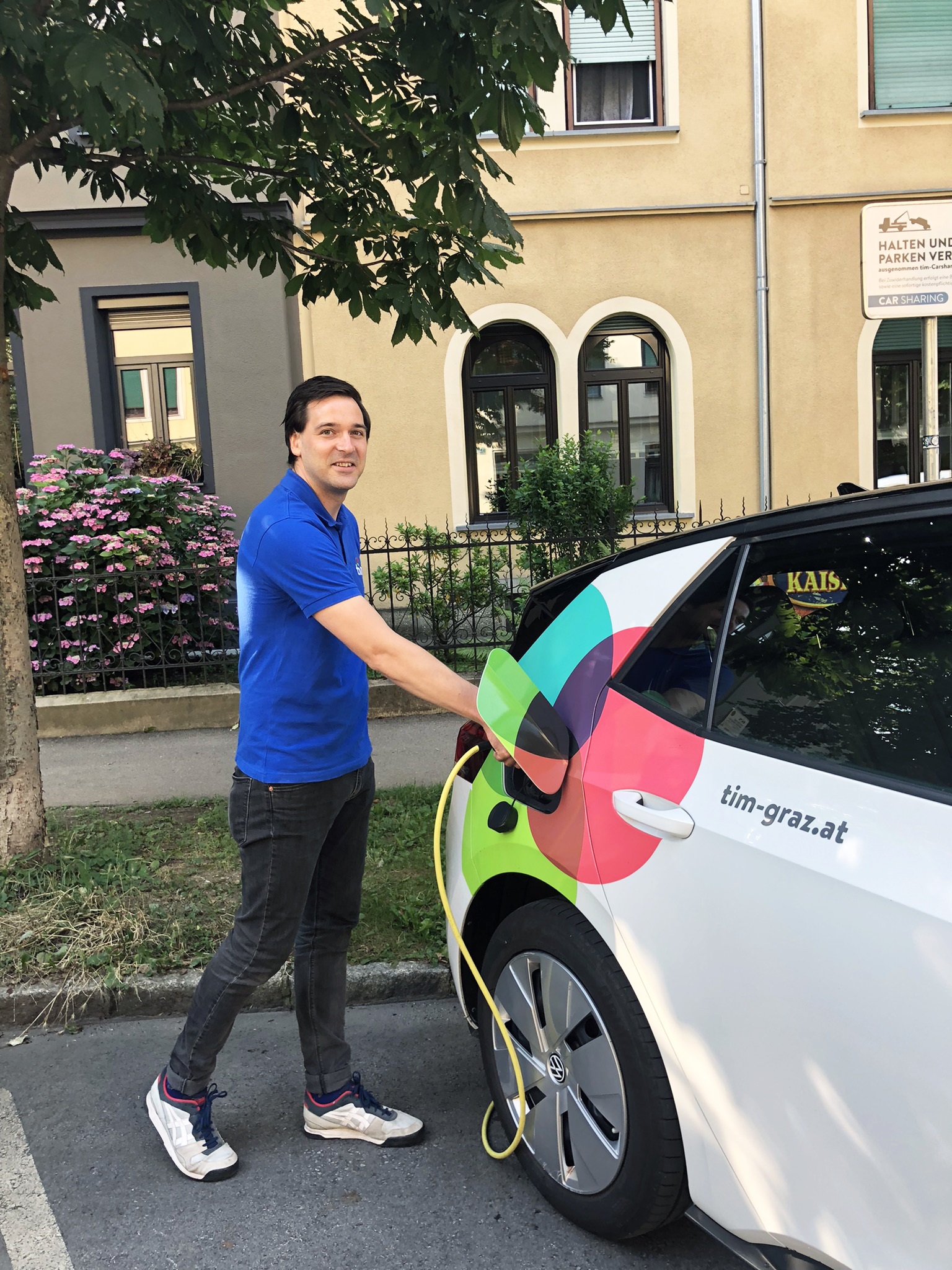 Mann lädt ein buntes E-Auto von tim-graz.at an einer Ladestation in Wohngegend auf.