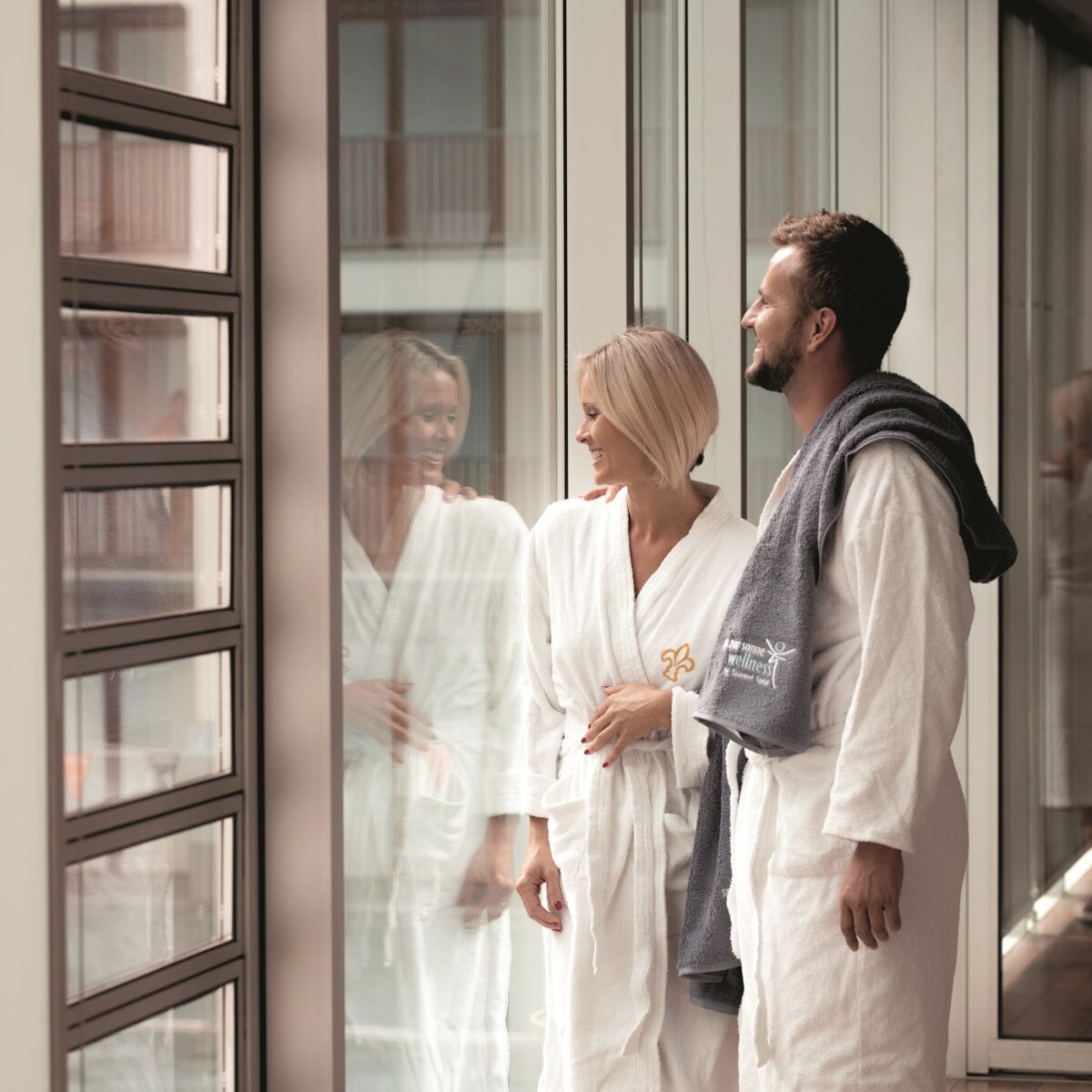Paar in Bademänteln steht lächelnd vor einer Glasfront in einem Wellnessbereich.