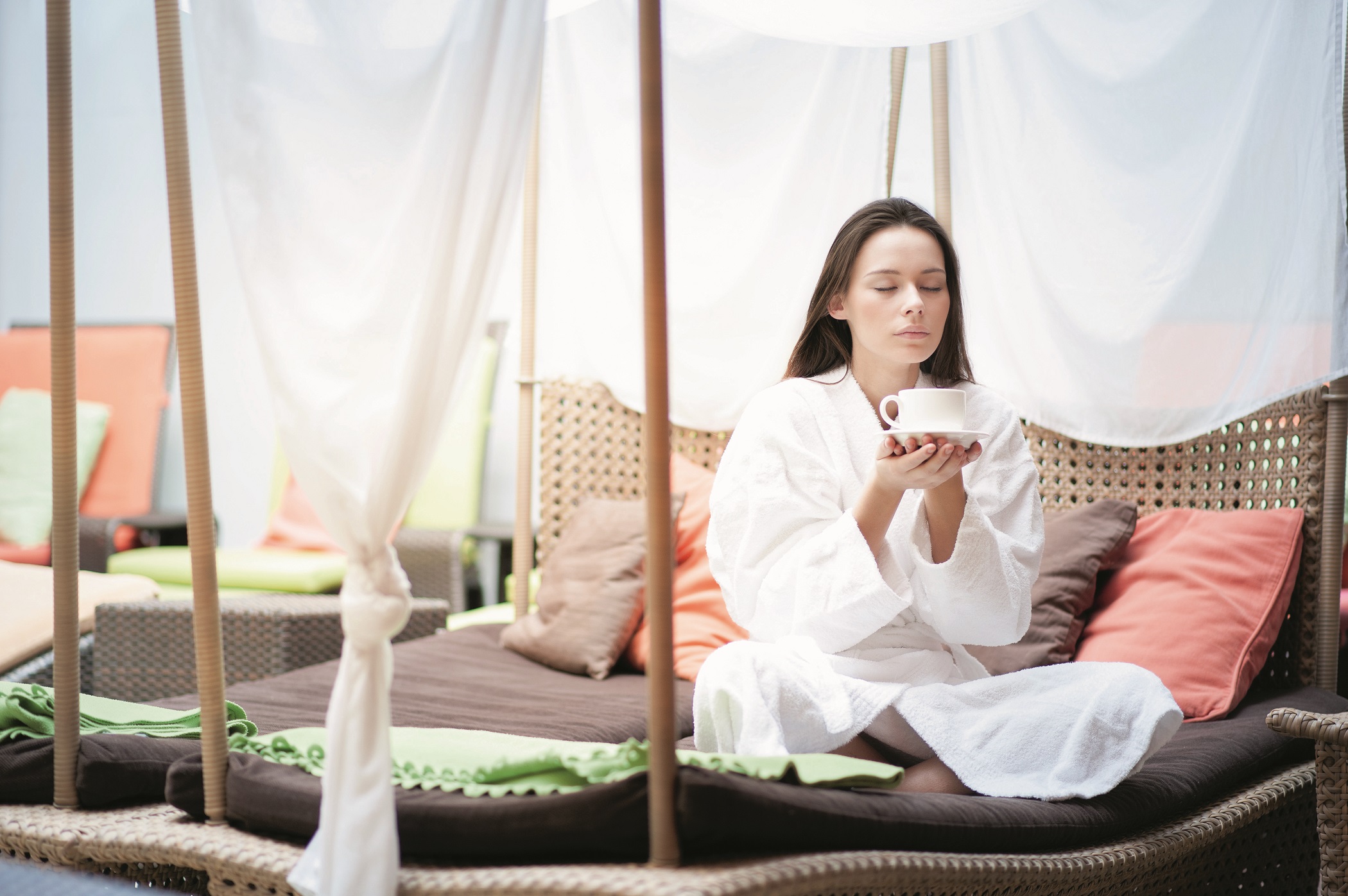 Frau in weißem Bademantel entspannt mit Tasse Tee in einer gemütlichen Lounge mit Kissen.