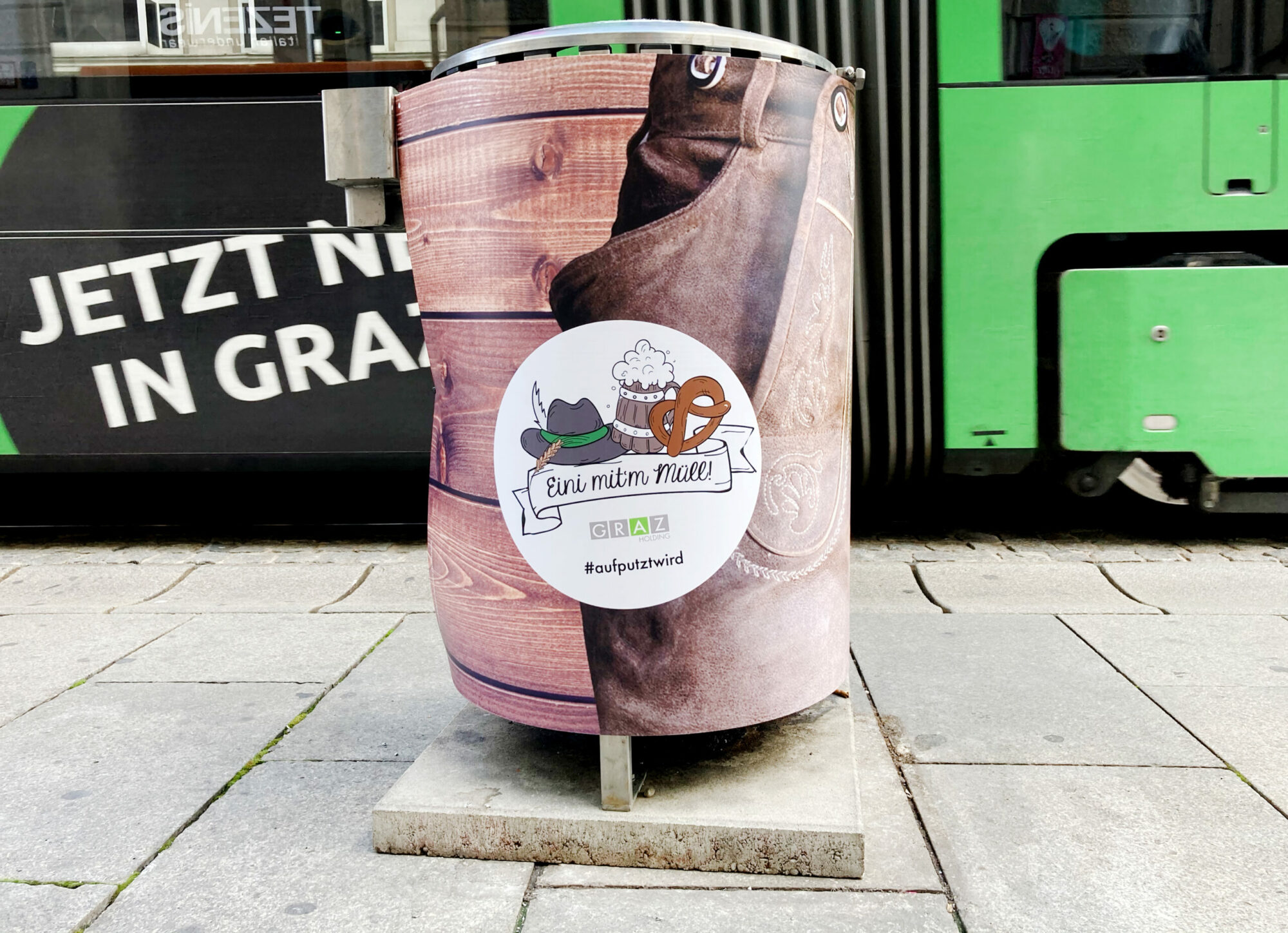 Mülleimer in Graz mit Holzdesign, Trachtenmotiv und Aufschrift 'Eini mit'm Müll!' vor Straßenbahn.