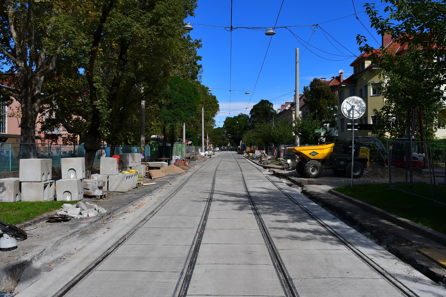 Straßenbauarbeiten an Gleisen, umgeben von Bäumen, Baustellenmaterialien und Baumaschinen.
