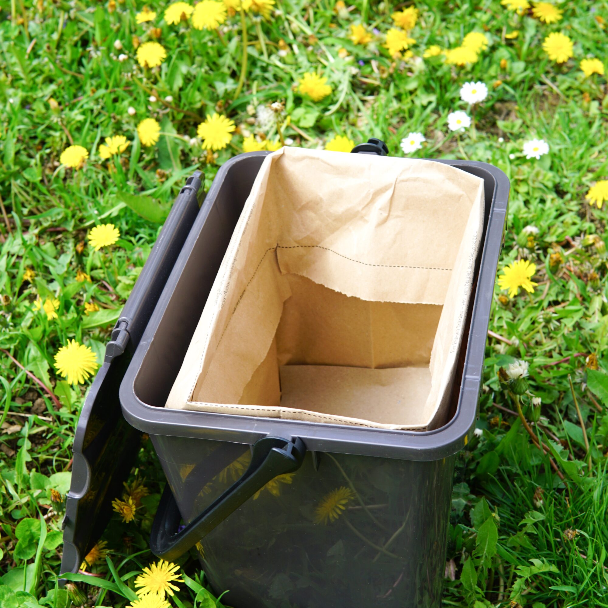 A black compost bin with a brown paper bag inside, sitting on a grassy meadow with yellow dandelions.