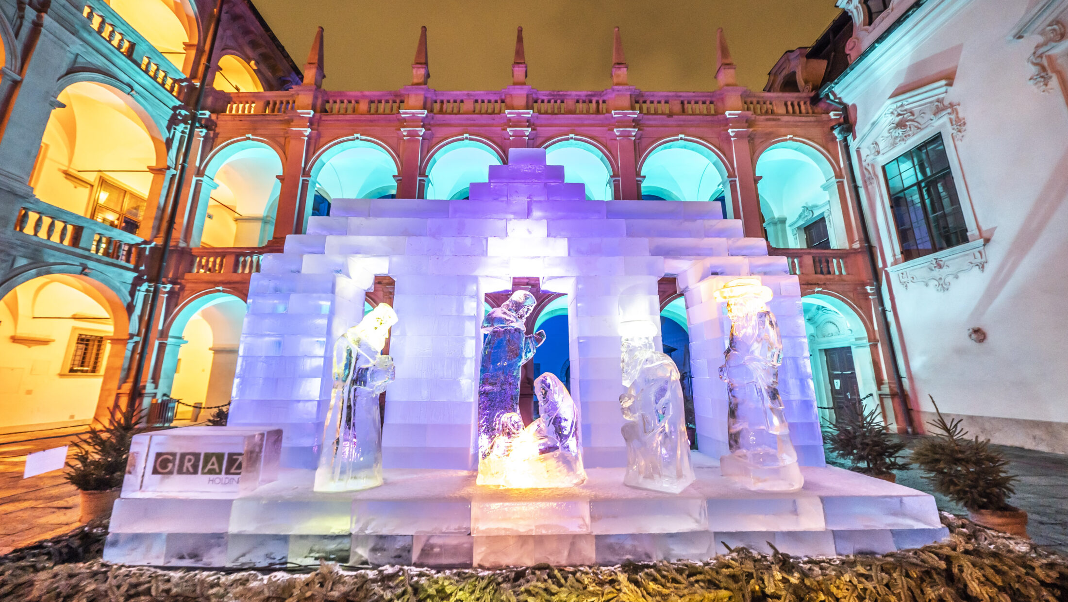 An illuminated ice sculpture depicting the Nativity scene in Graz, Austria. The sculpture is lit with colorful lights