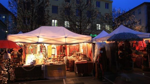 Standl am Adventmarkt am Tummelplatz . Es ist Abends und in der Auslage eines Standls sind verschiedenen Stoffe, Kleidungsstücke und Schmuck zu sehen.
