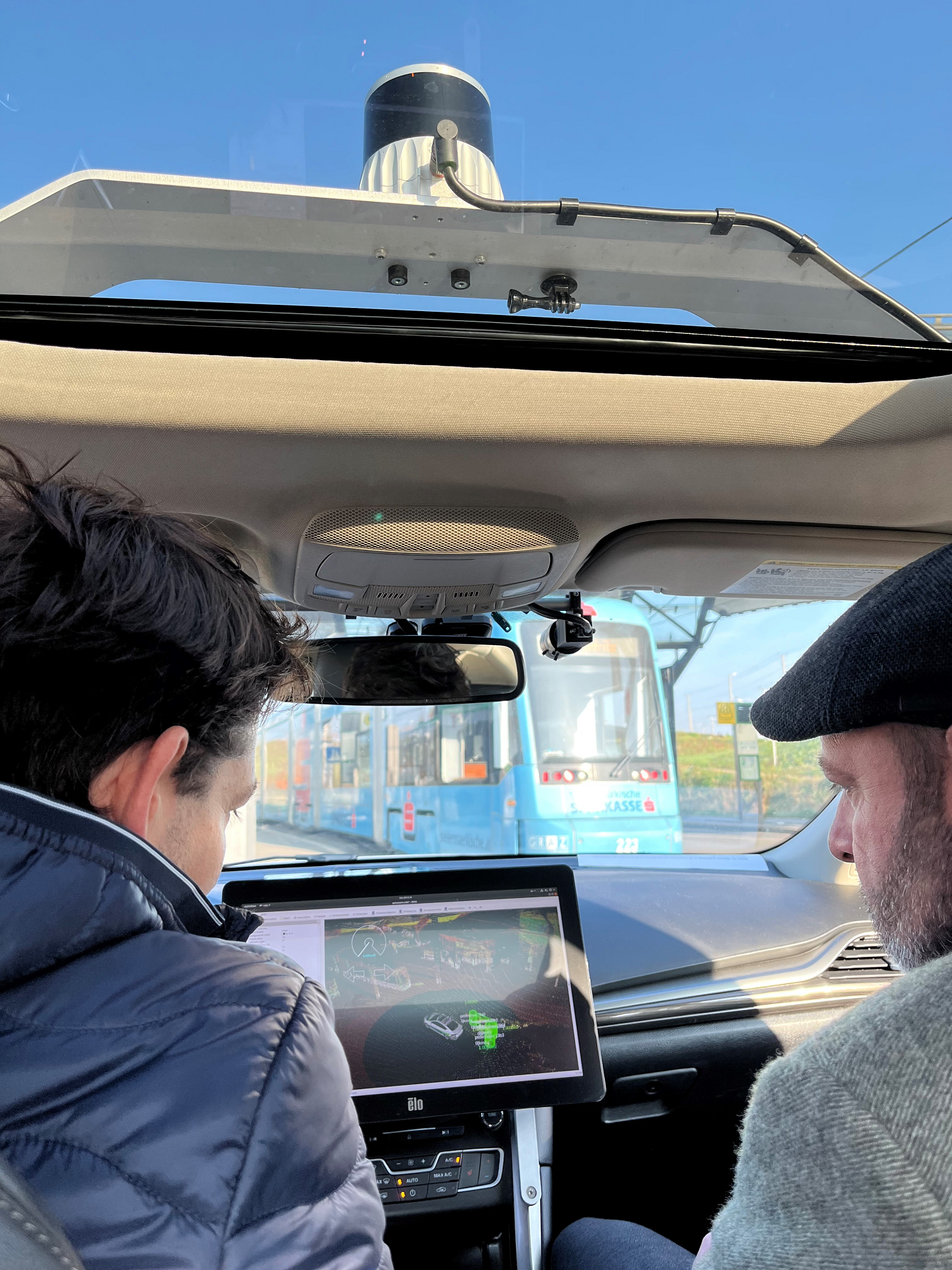 Zwei Personen im Auto mit Blick auf Monitor und Sensoren, blauer Himmel und Straßenbahn dahinter