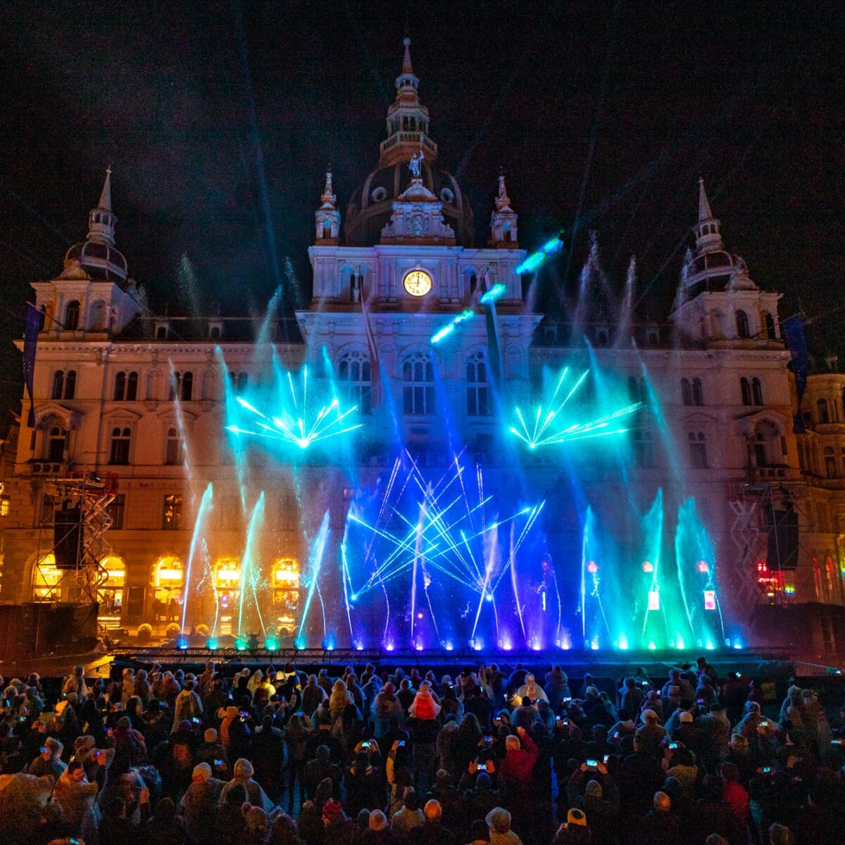 Lichtspektakel vor dem Grazer Rathaus mit bunten Wasserfontänen und Menschenmenge bei Nacht.