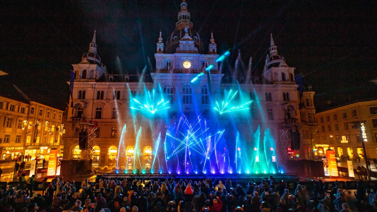 Lichtspektakel vor dem Grazer Rathaus mit bunten Wasserfontänen und Menschenmenge bei Nacht.