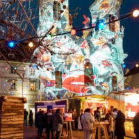 Adventmarkt am Mariahilferplatz mit bunter Beleuchtung und Projektionen auf die Kirche.