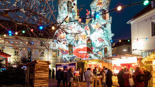 Adventmarkt am Mariahilferplatz mit bunter Beleuchtung und Projektionen auf die Kirche.